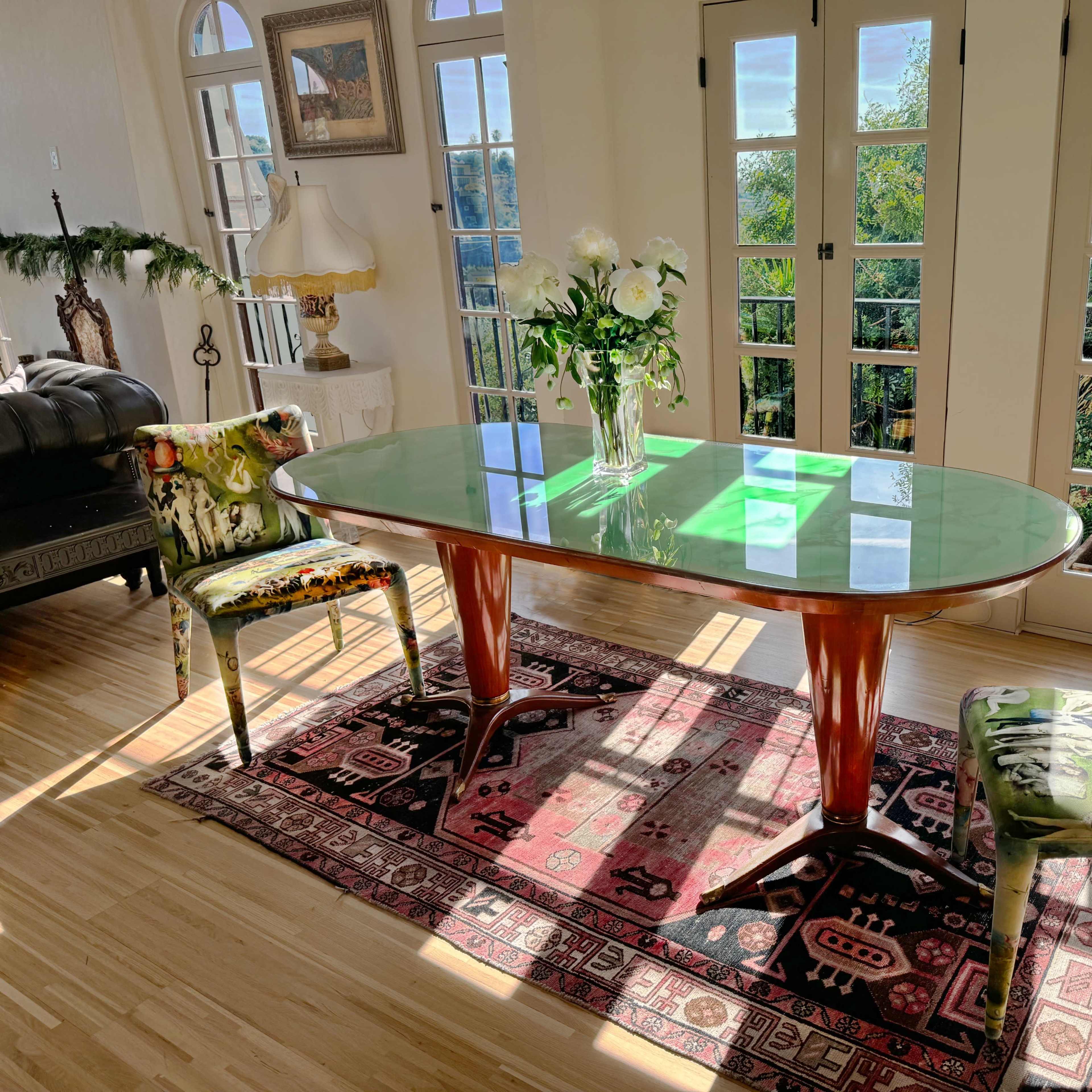 A dining table with a green glass top is surrounded by two patterned chairs on a colorful rug, with large windows allowing sunlight to enter the room.