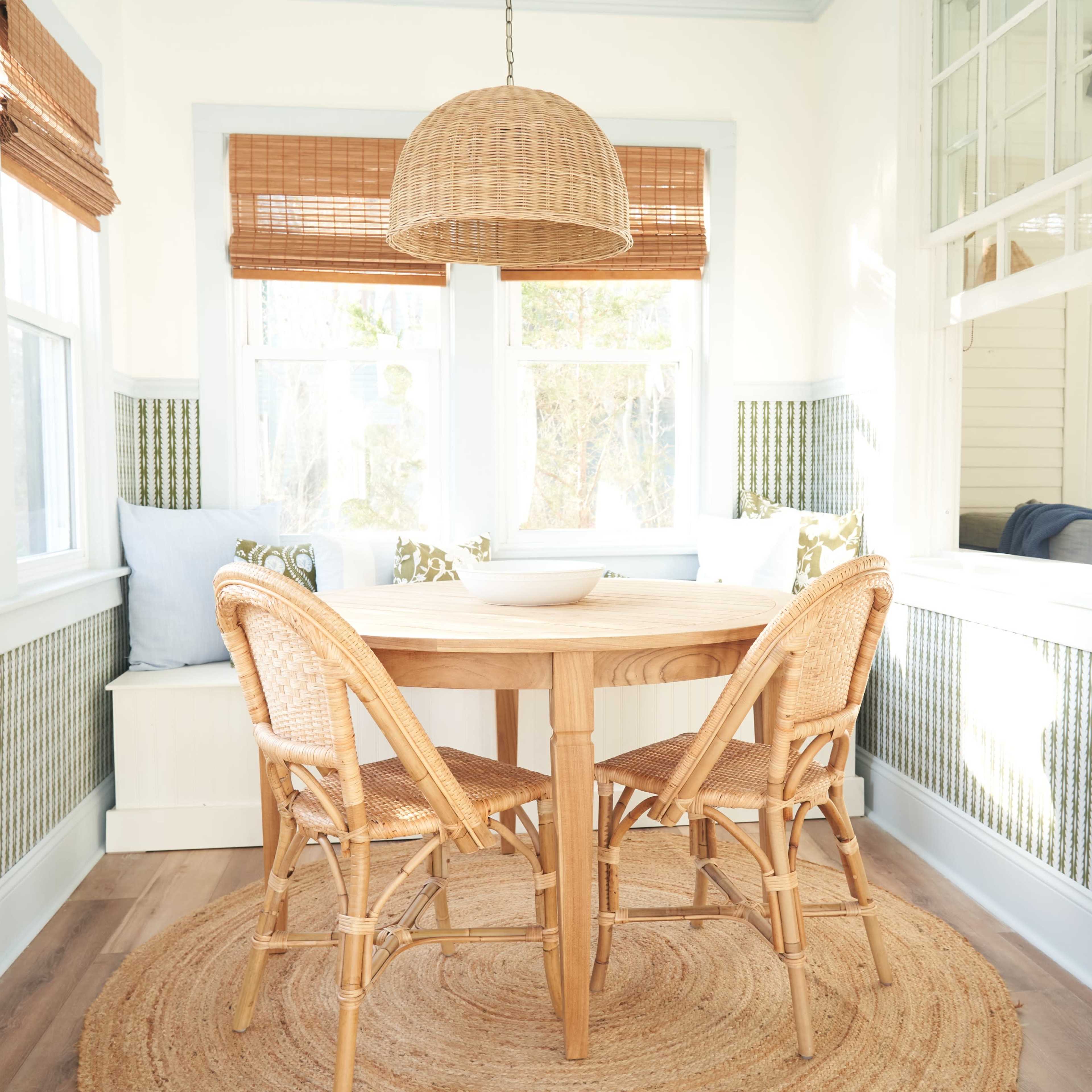 A round wooden table with four wicker chairs is centered in a sunlit dining area featuring large windows and patterned wallpaper.