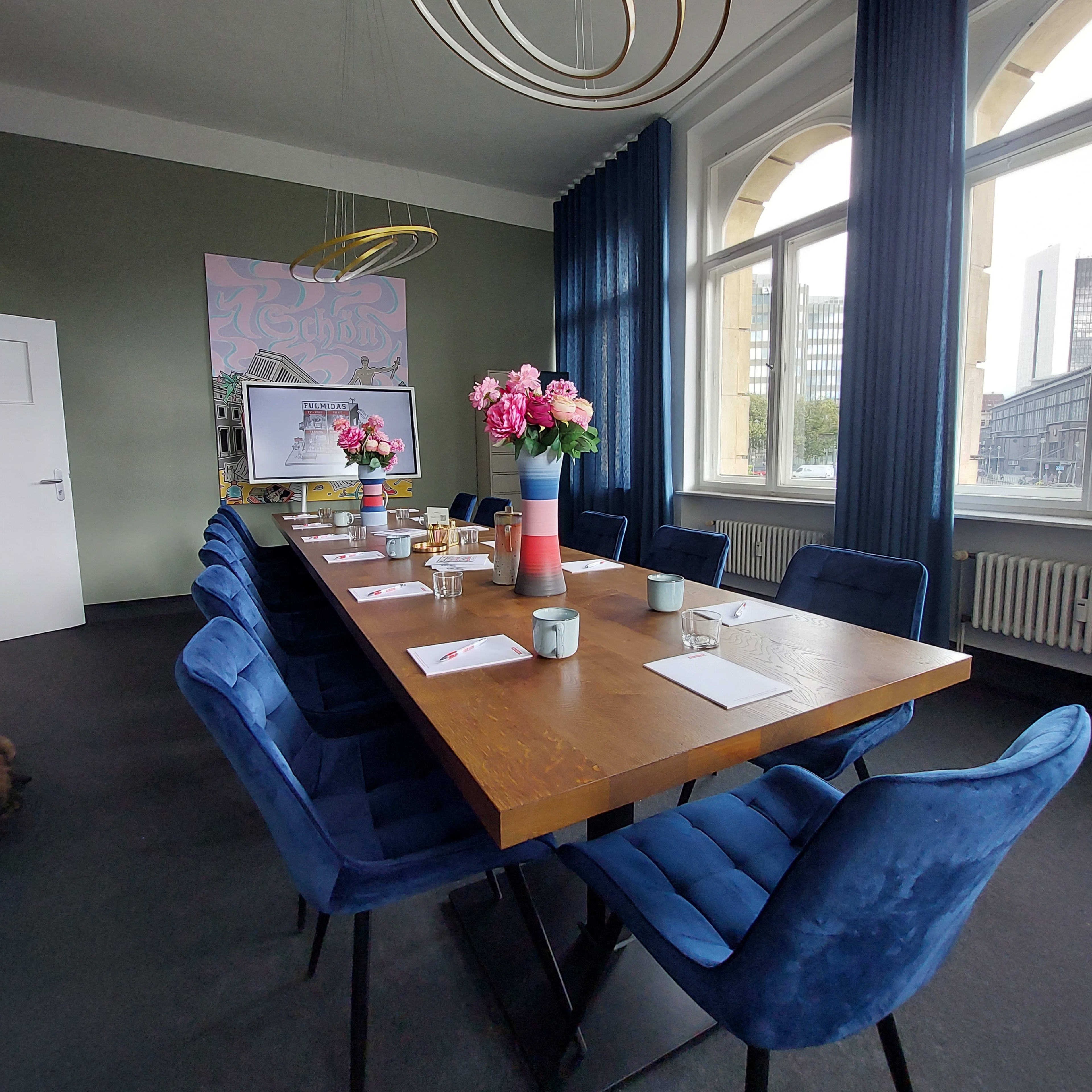 A conference room features a long wooden table surrounded by blue chairs, with floral centerpieces and large windows draped with curtains.