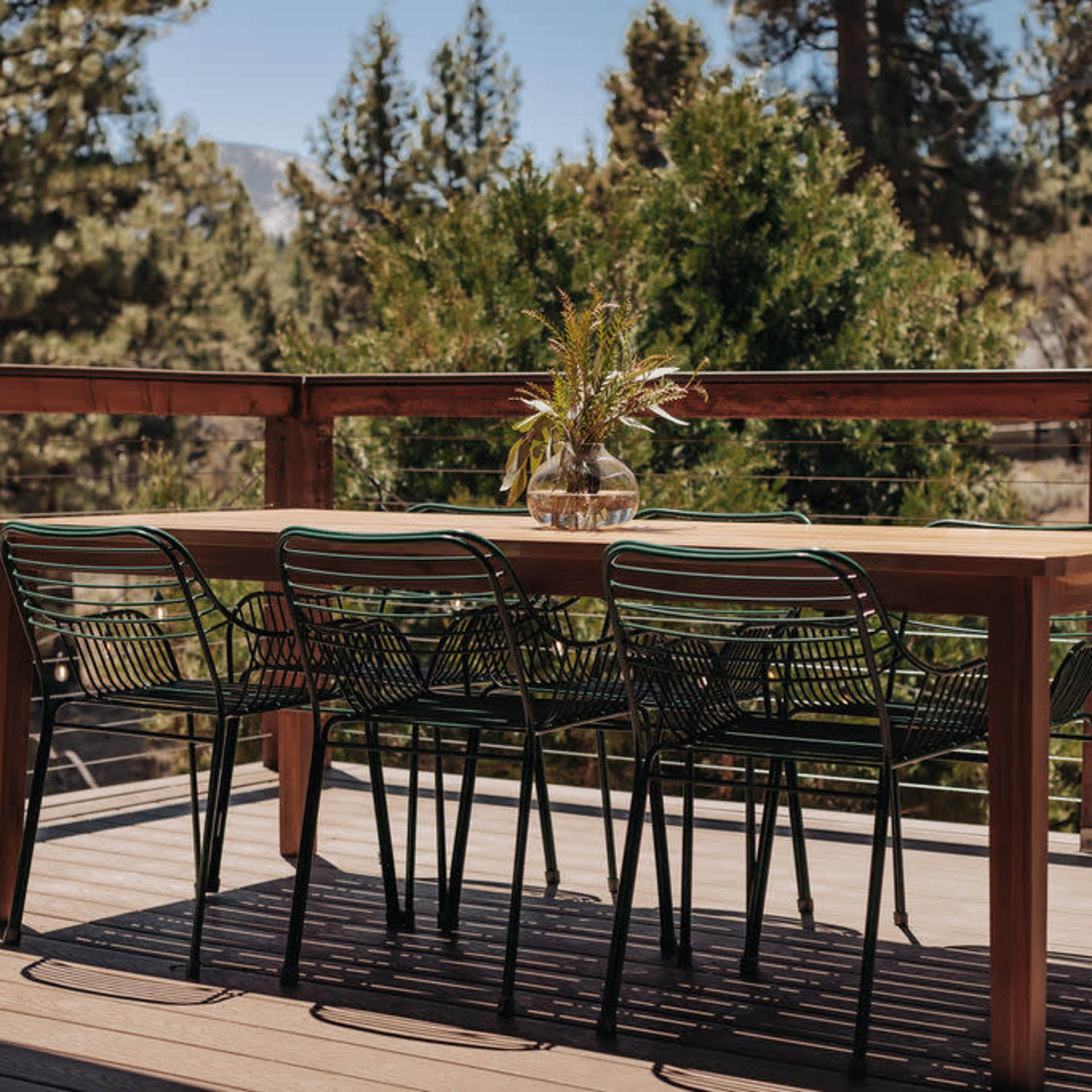 A wooden dining table with six metal chairs is set on a deck surrounded by tall trees and a clear blue sky.