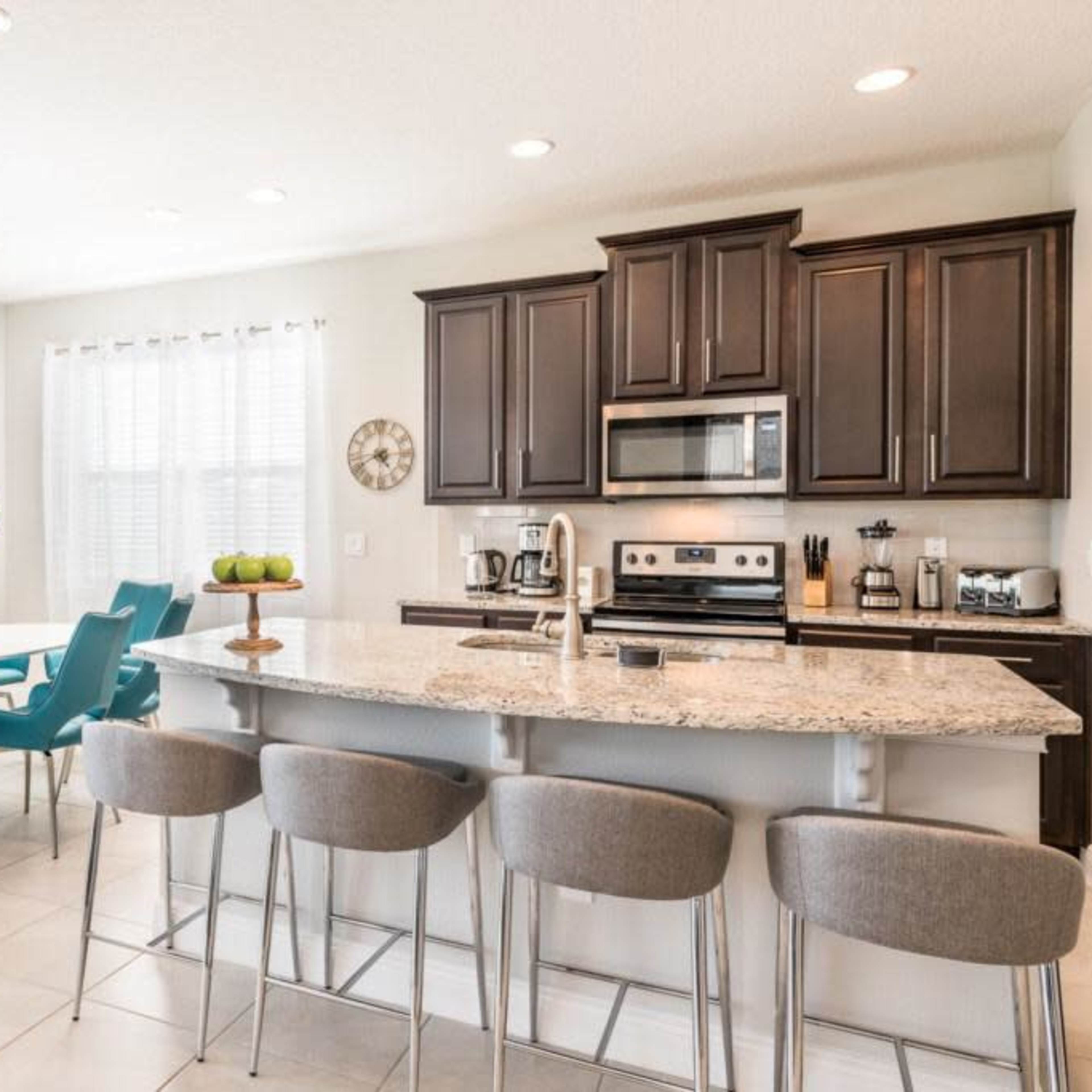 A modern kitchen features dark cabinets, a granite island with gray bar stools, and a dining area with a large table and teal chairs.