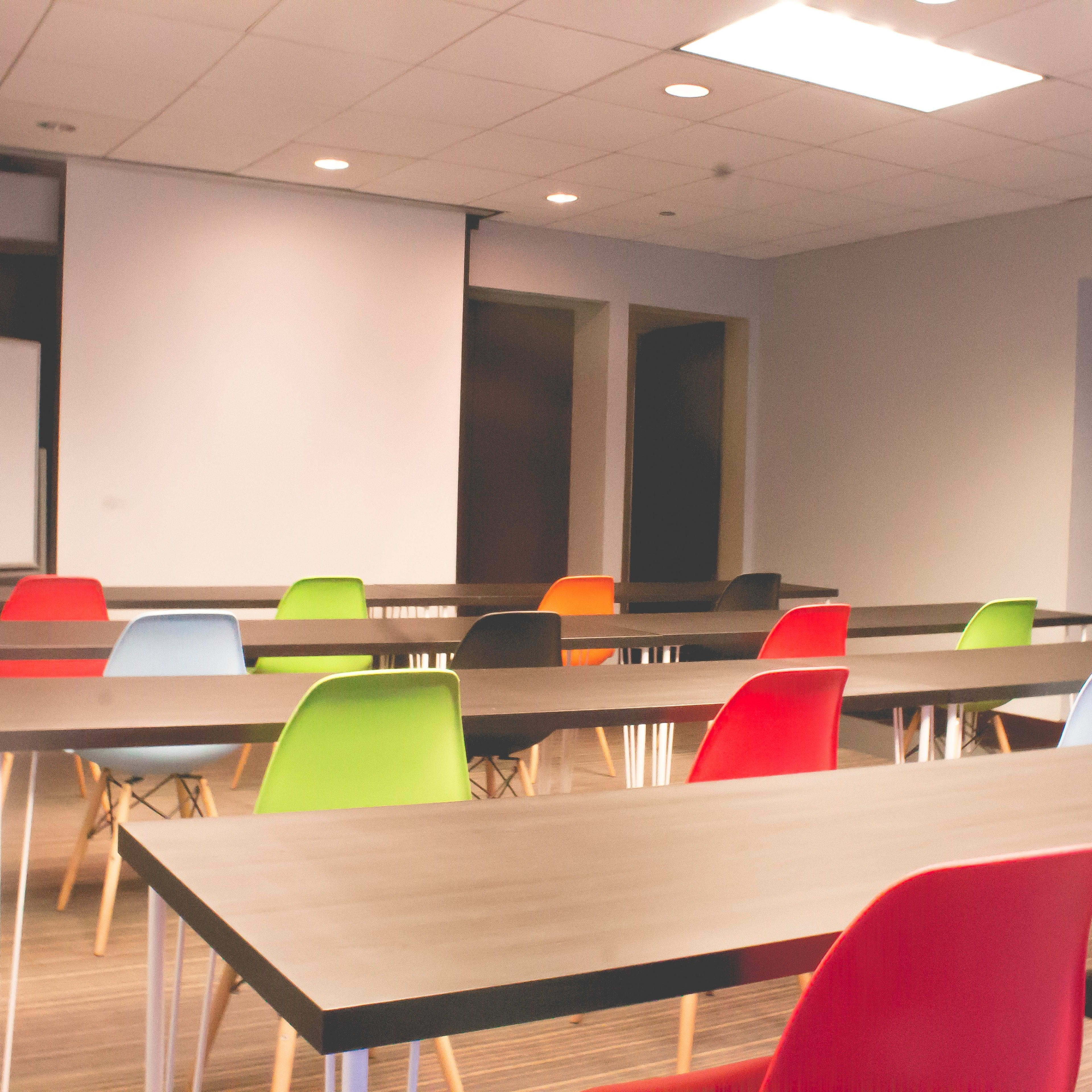 A modern conference room features several tables arranged in rows, with colorful chairs and a projector screen at the front.