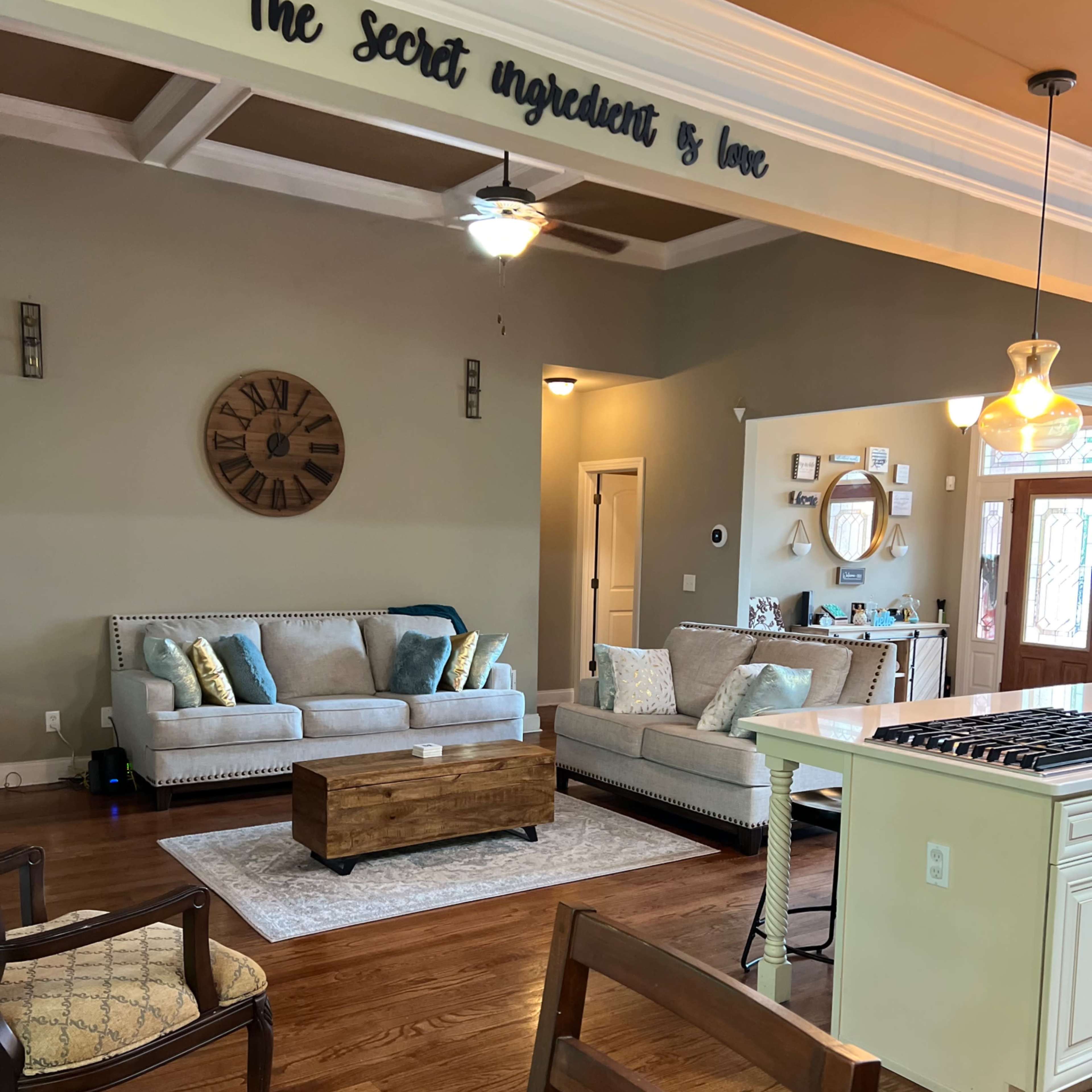 A living room and kitchen area featuring a beige sofa, wooden coffee table, and wall decor with the phrase "The secret ingredient is love."