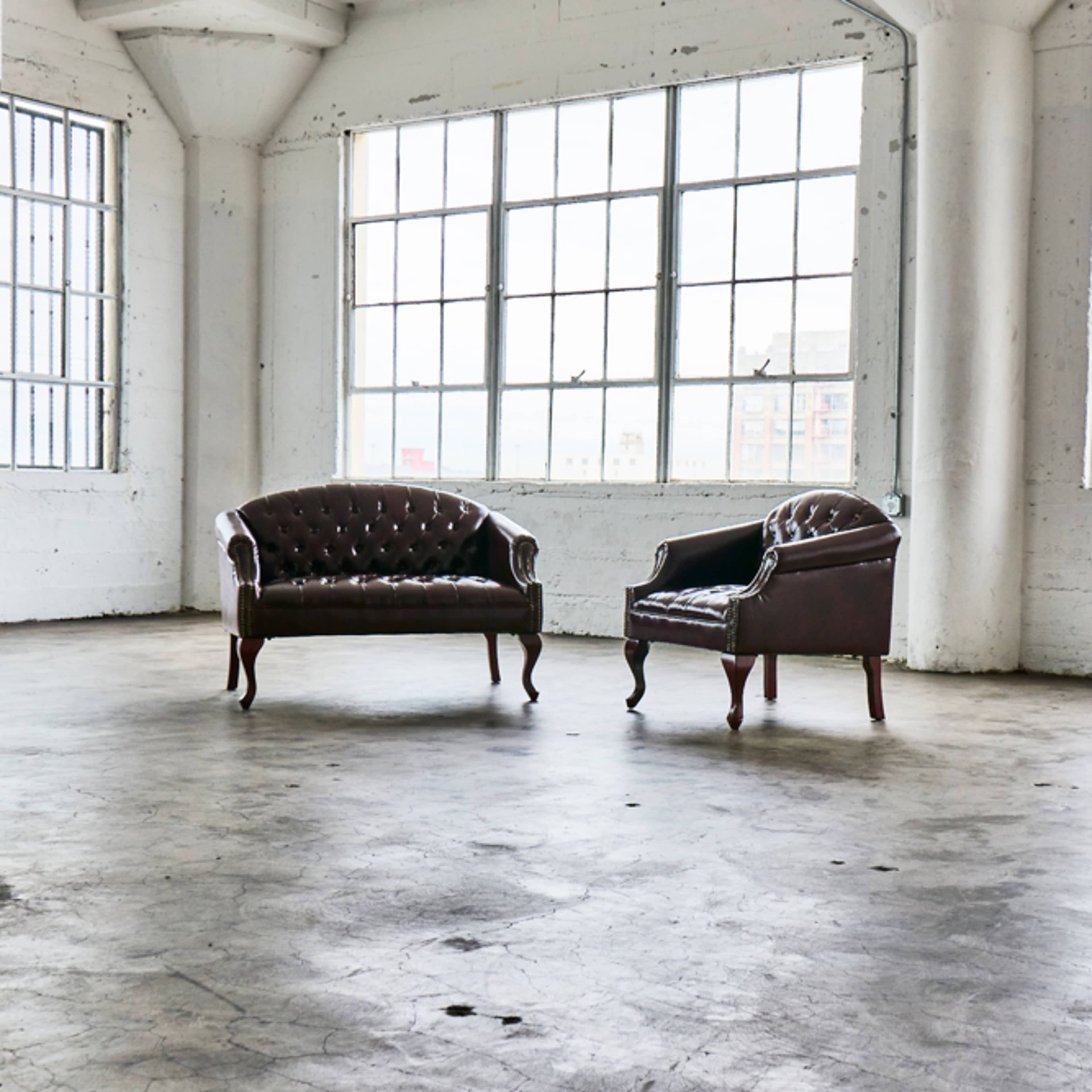 Two leather chairs are positioned on a concrete floor in a spacious room with large windows.