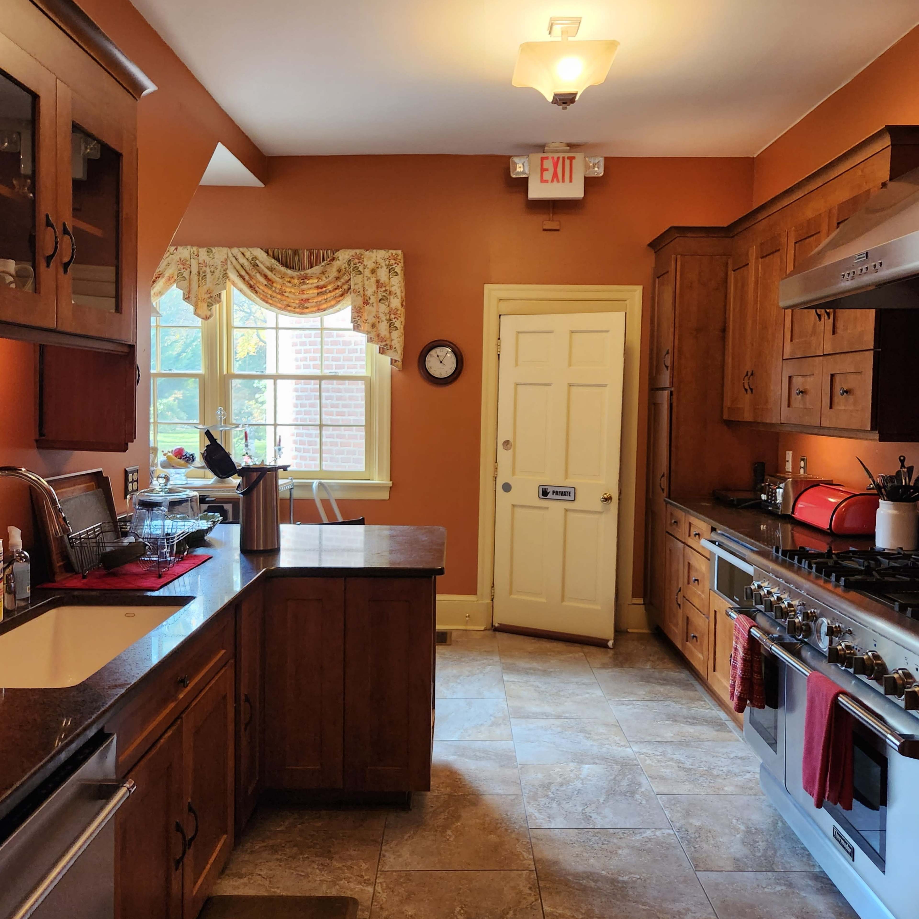 A kitchen with wooden cabinetry and granite countertops features a stove, a window with floral curtains, and a door leading to another room.