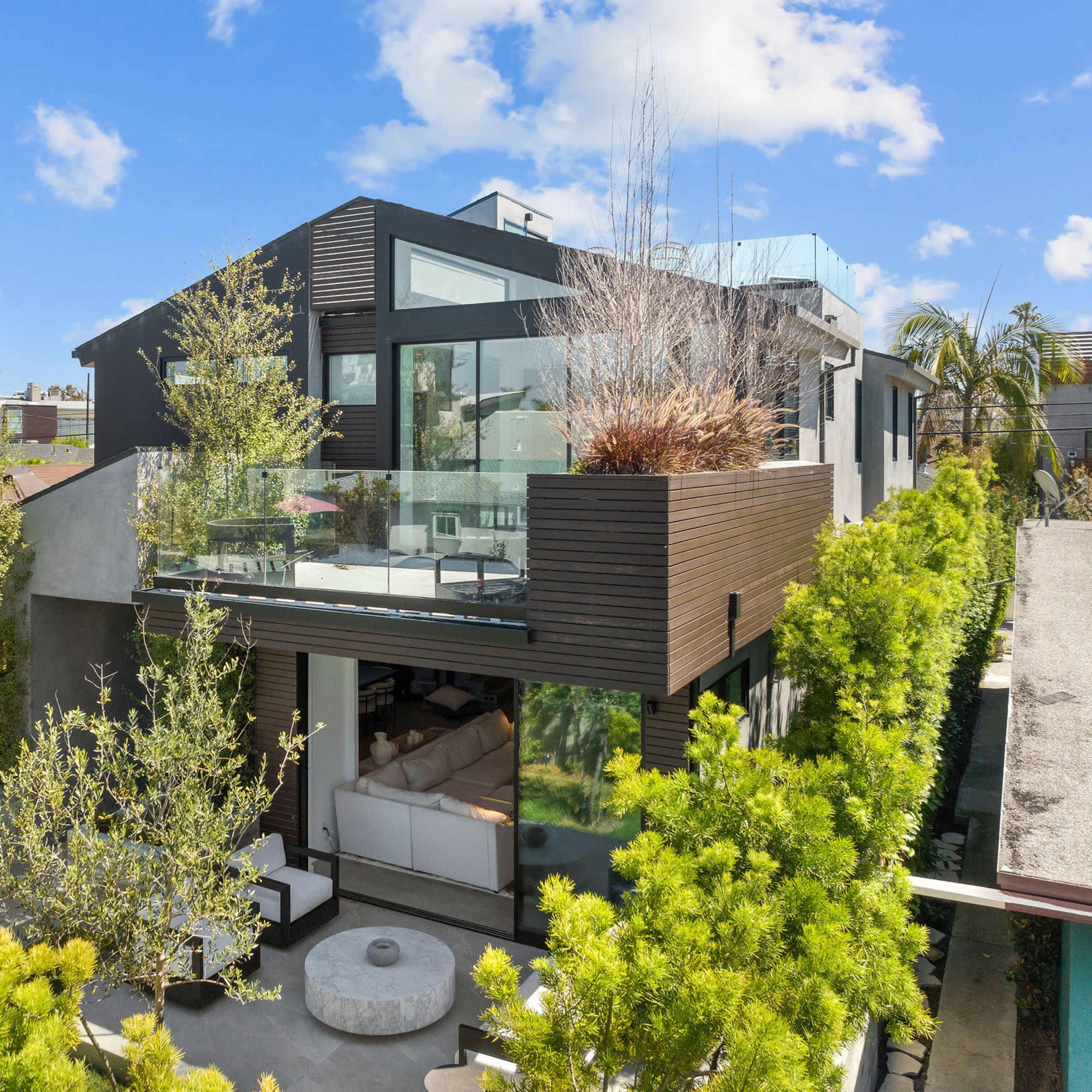A modern two-story house features large glass windows and a spacious balcony surrounded by greenery.