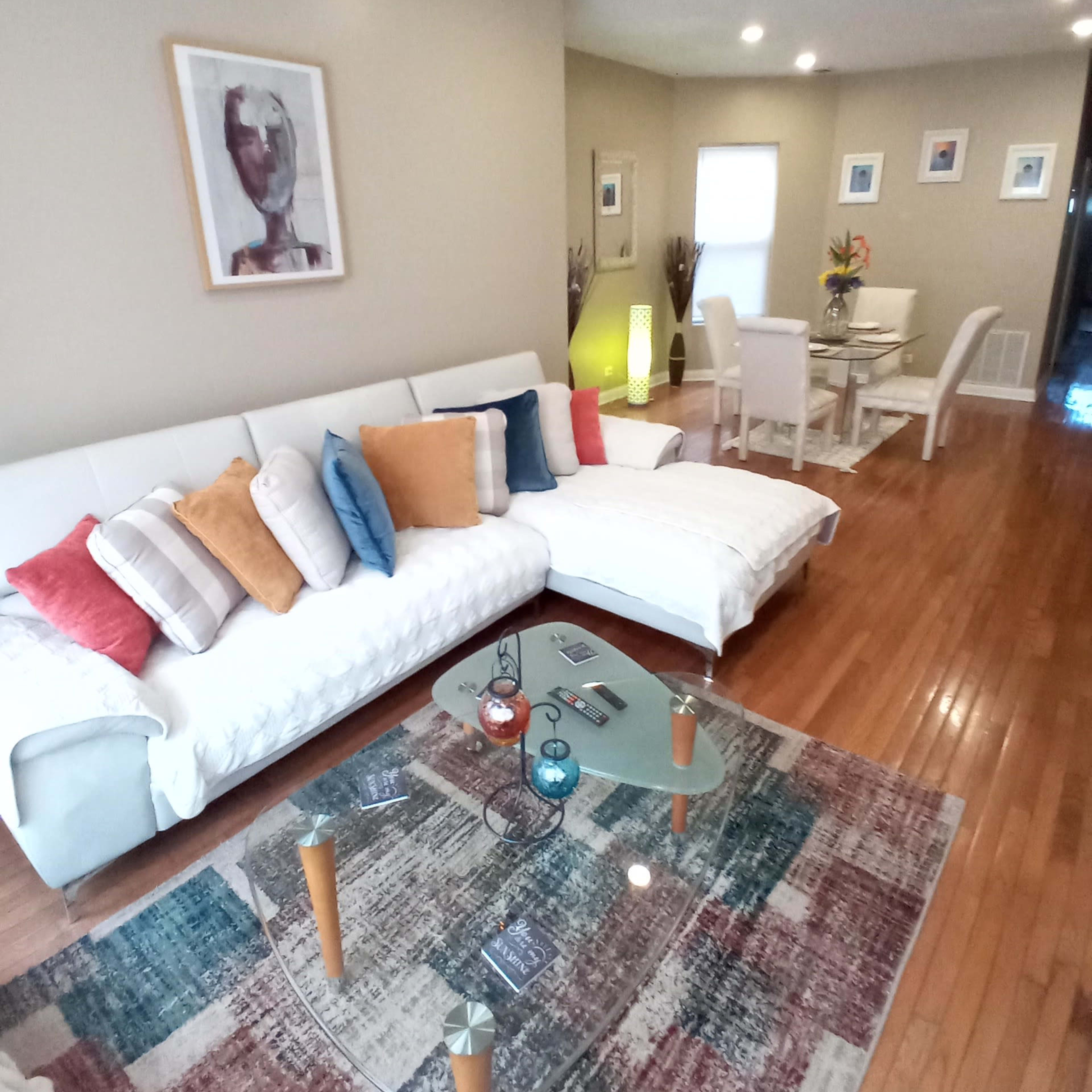 A living room with a white sectional sofa, a glass coffee table, and a dining area visible in the background.