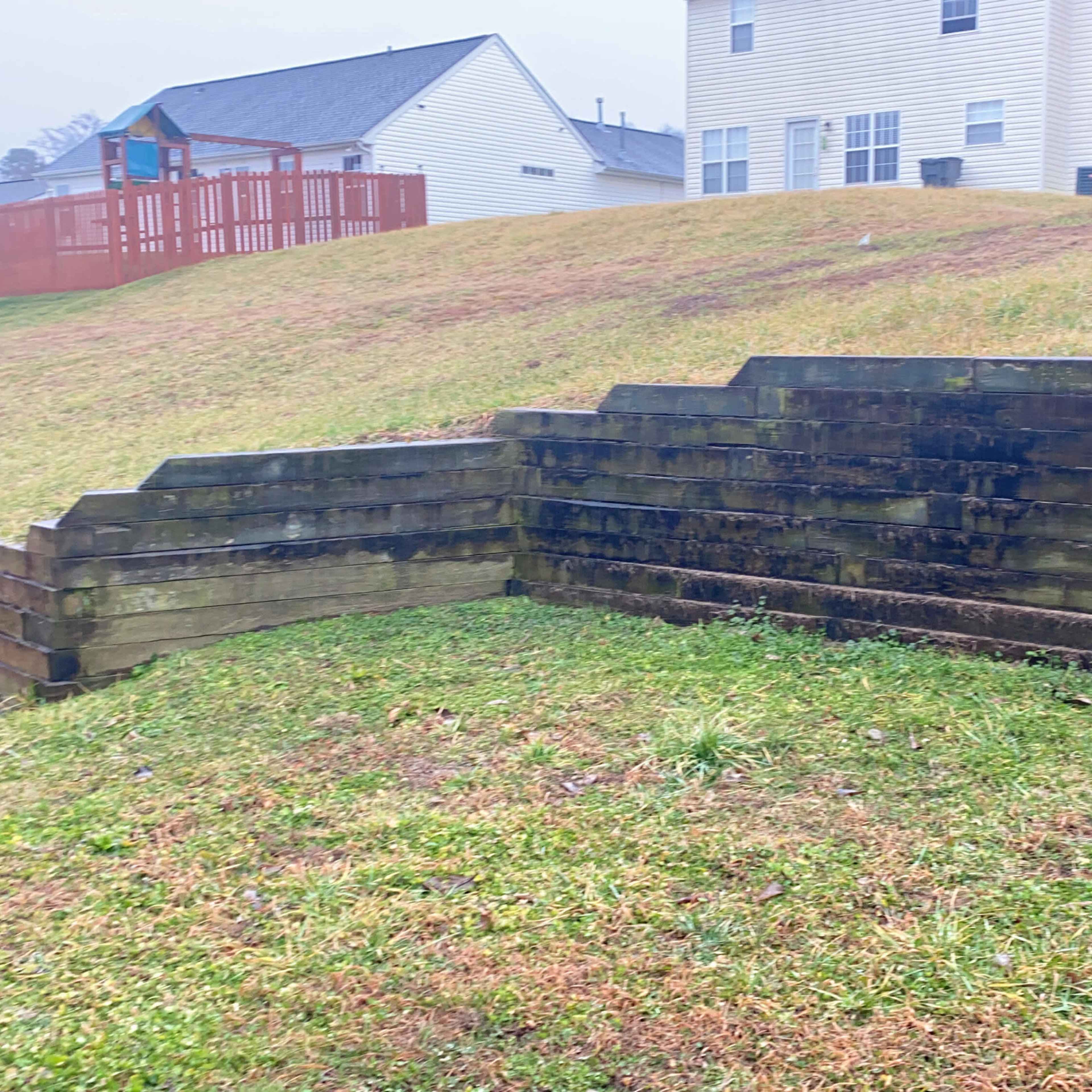 A grassy area with a sloped hill and a wooden retaining wall partially covered in moss, bordered by two residential buildings.