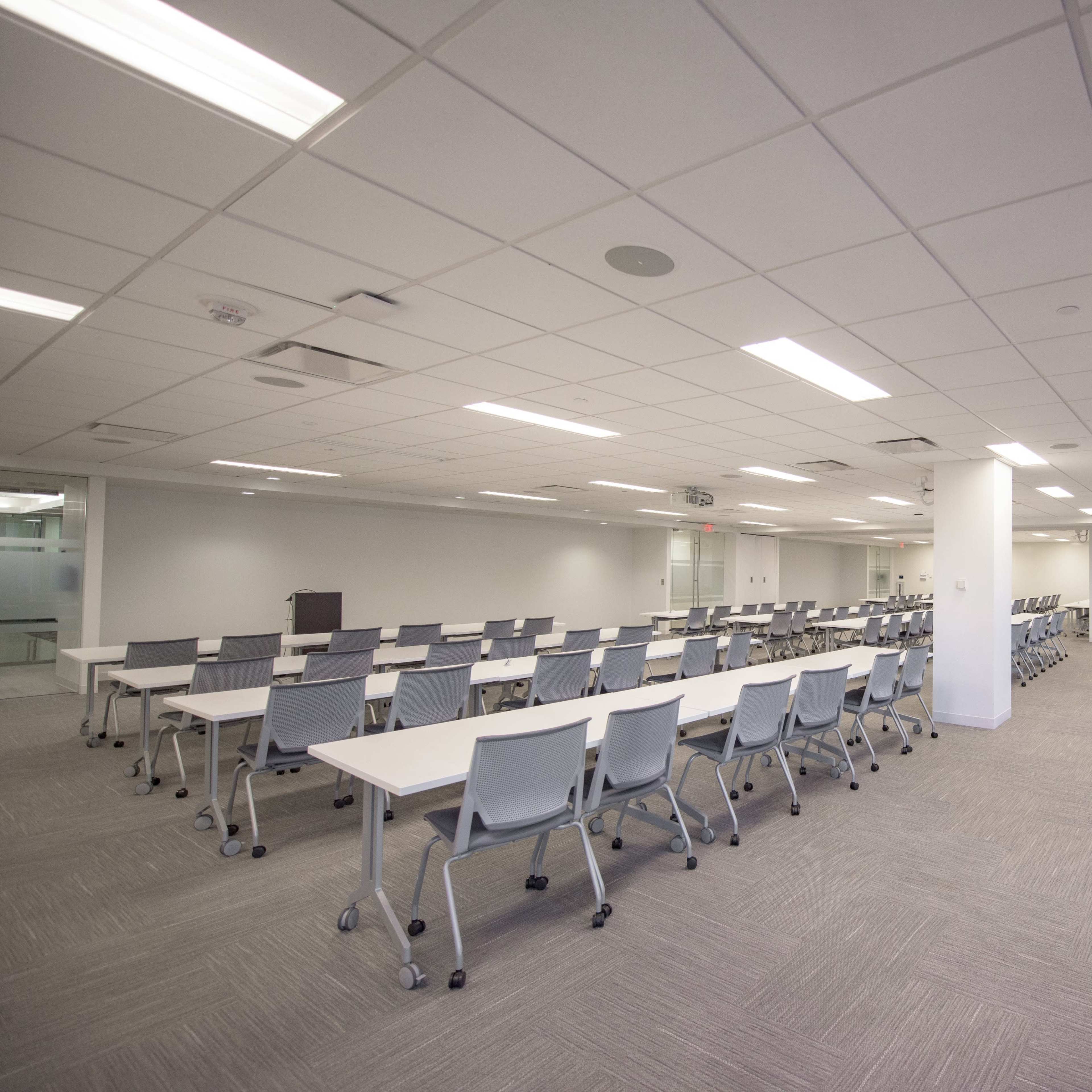 A spacious meeting room features rows of white tables and chairs, with large windows and neutral-colored carpeting.