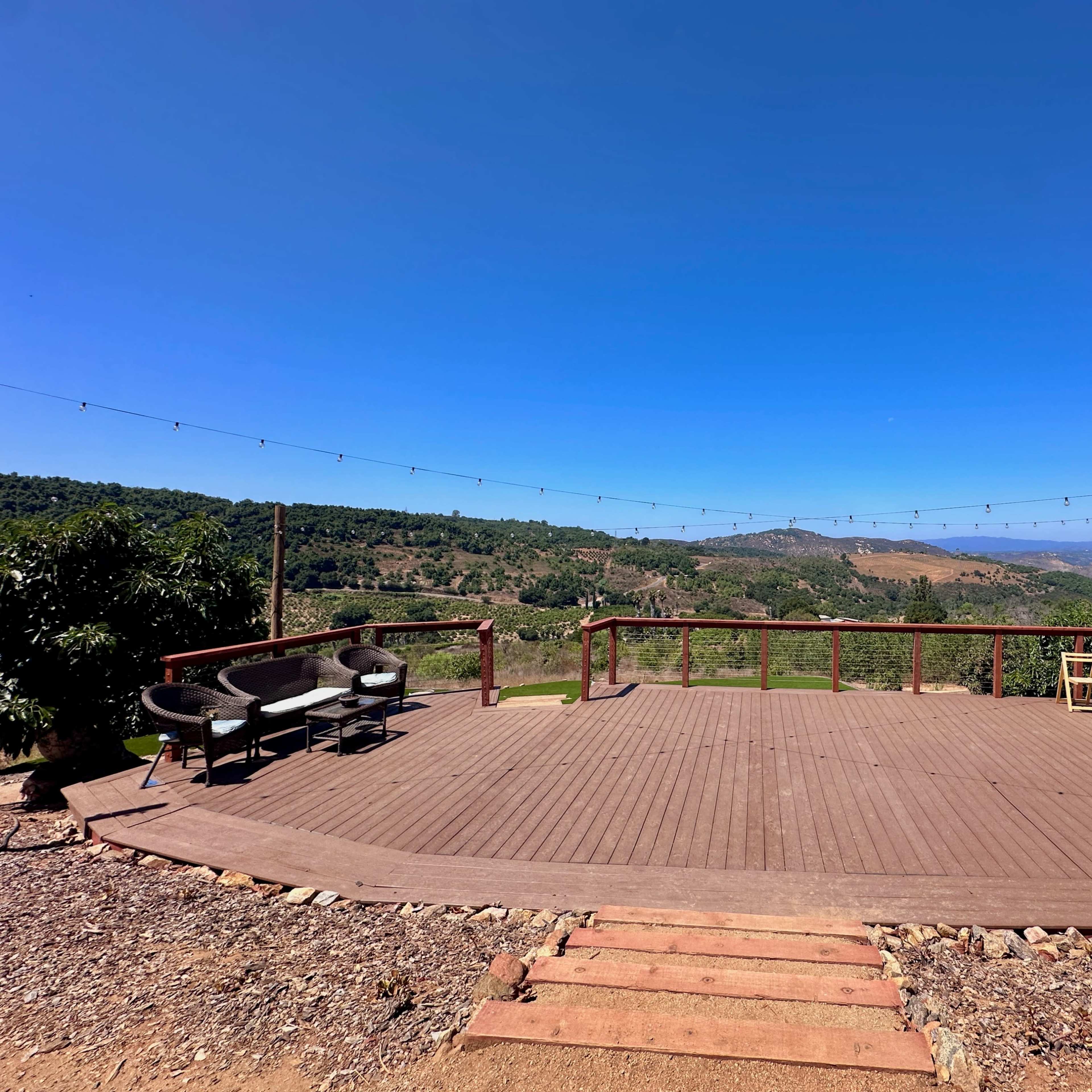 An empty wooden deck overlooks a lush valley under a clear blue sky, with string lights hanging above.