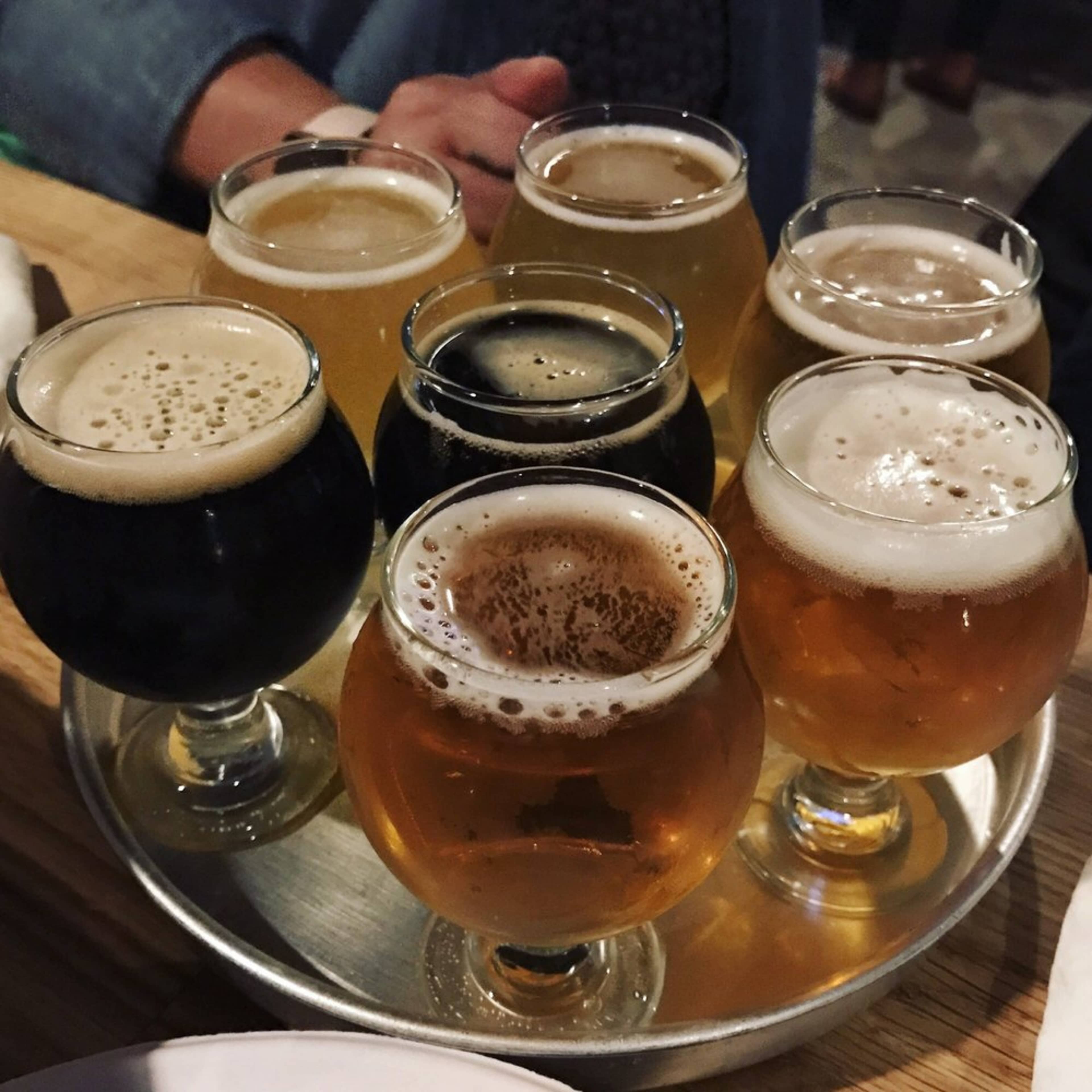 A tray holding eight glasses of different beers, arranged in a circle, with varying colors and foam levels.