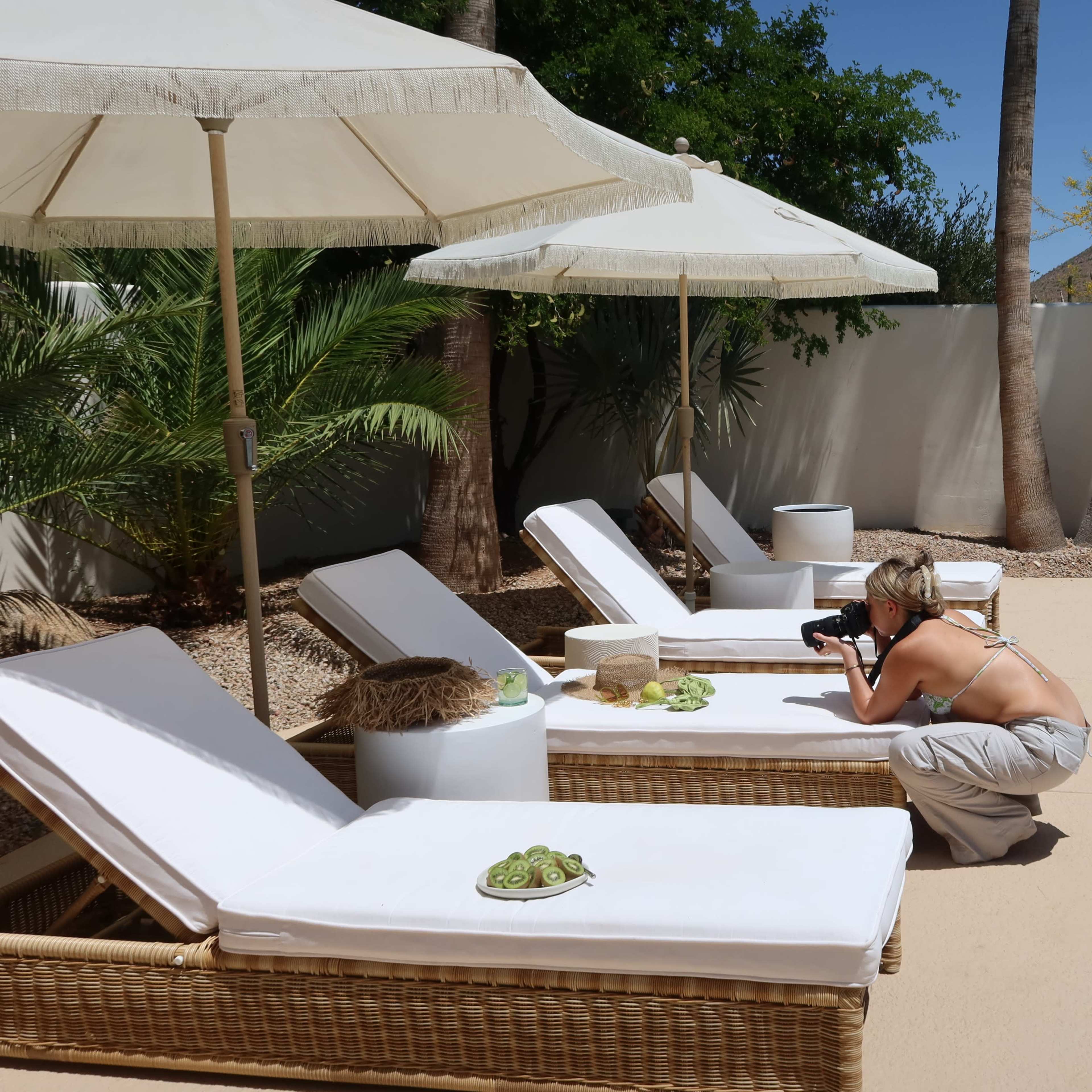 A woman crouches beside a pool, taking photos of a decorative arrangement on a lounge chair under an umbrella.