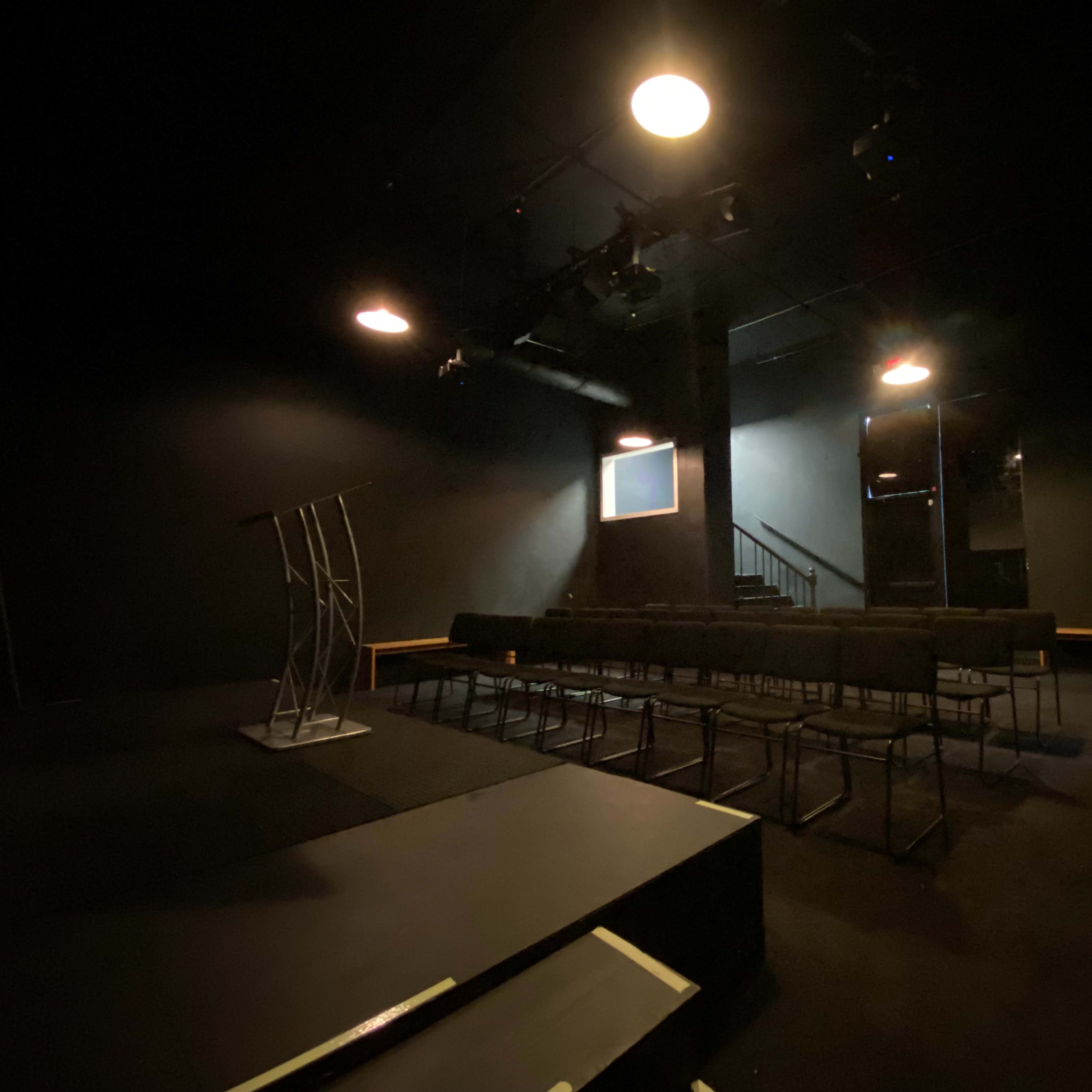 A dimly lit theater space with a podium, rows of chairs, and a staircase in the background.