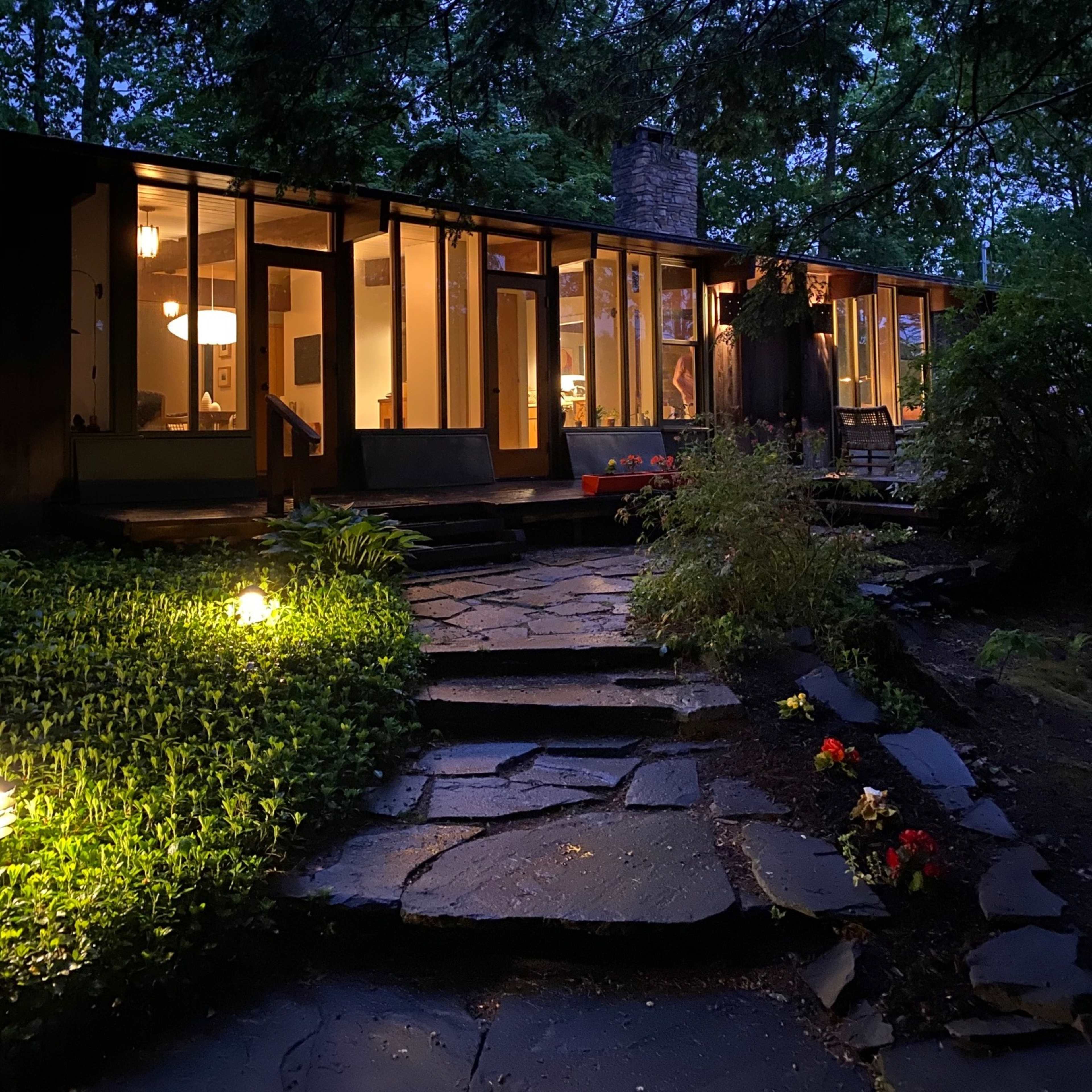 A stone pathway leads to a modern house illuminated in the twilight, surrounded by greenery and flowers.