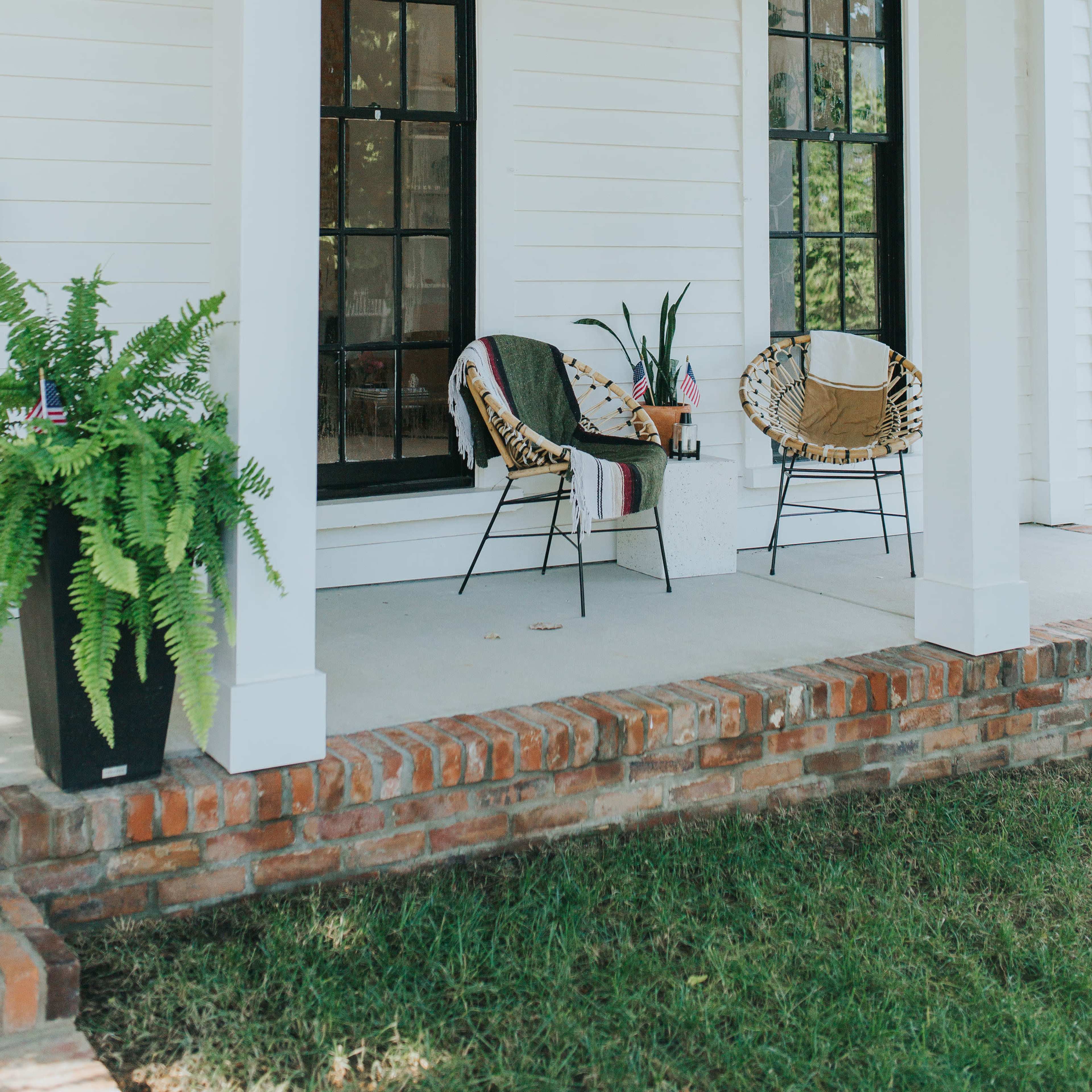 A porch with two patterned chairs, a potted plant, and a ceiling fan, surrounded by white walls and large windows.
