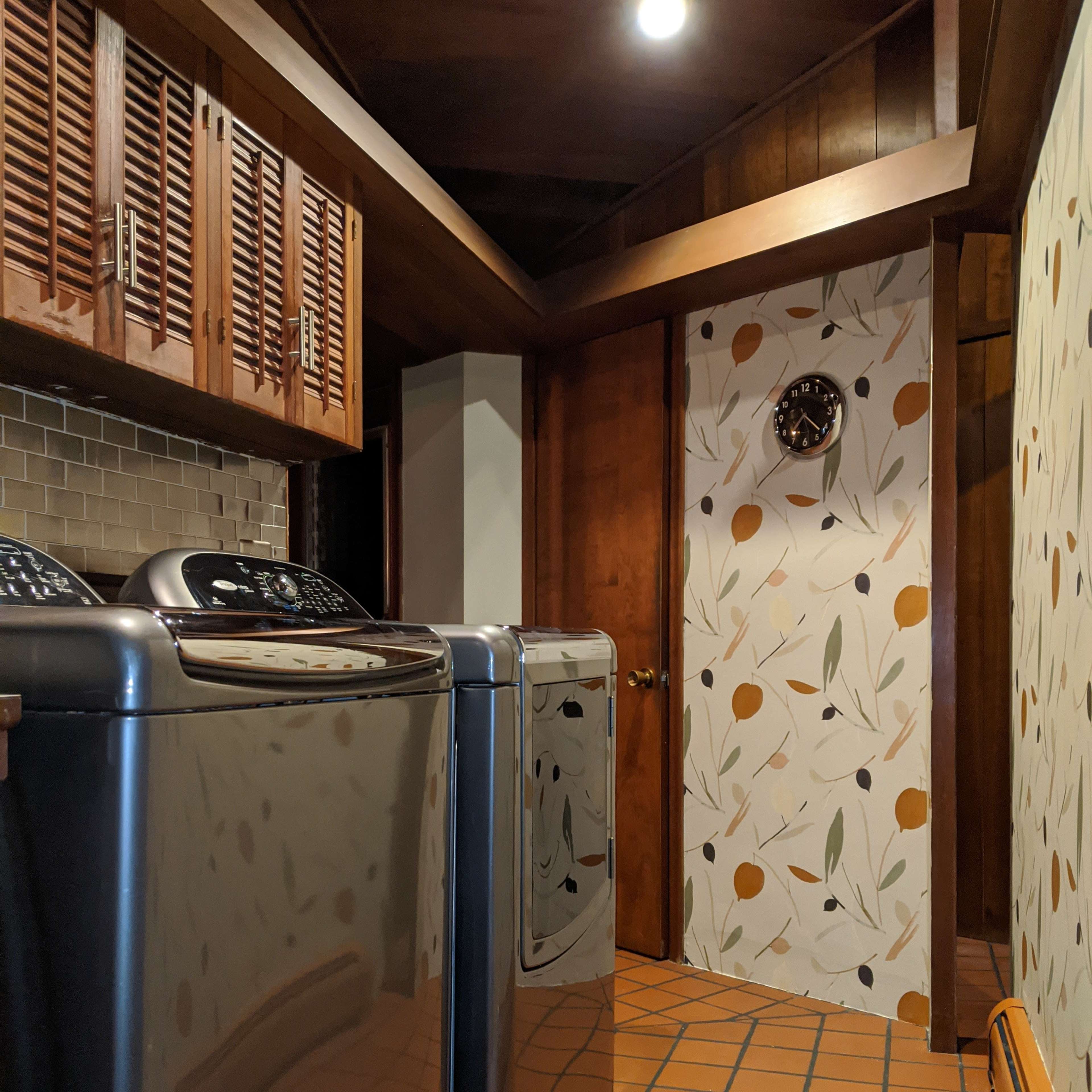A laundry room featuring two modern washing machines facing a decorative wall with a clock and patterned wallpaper.