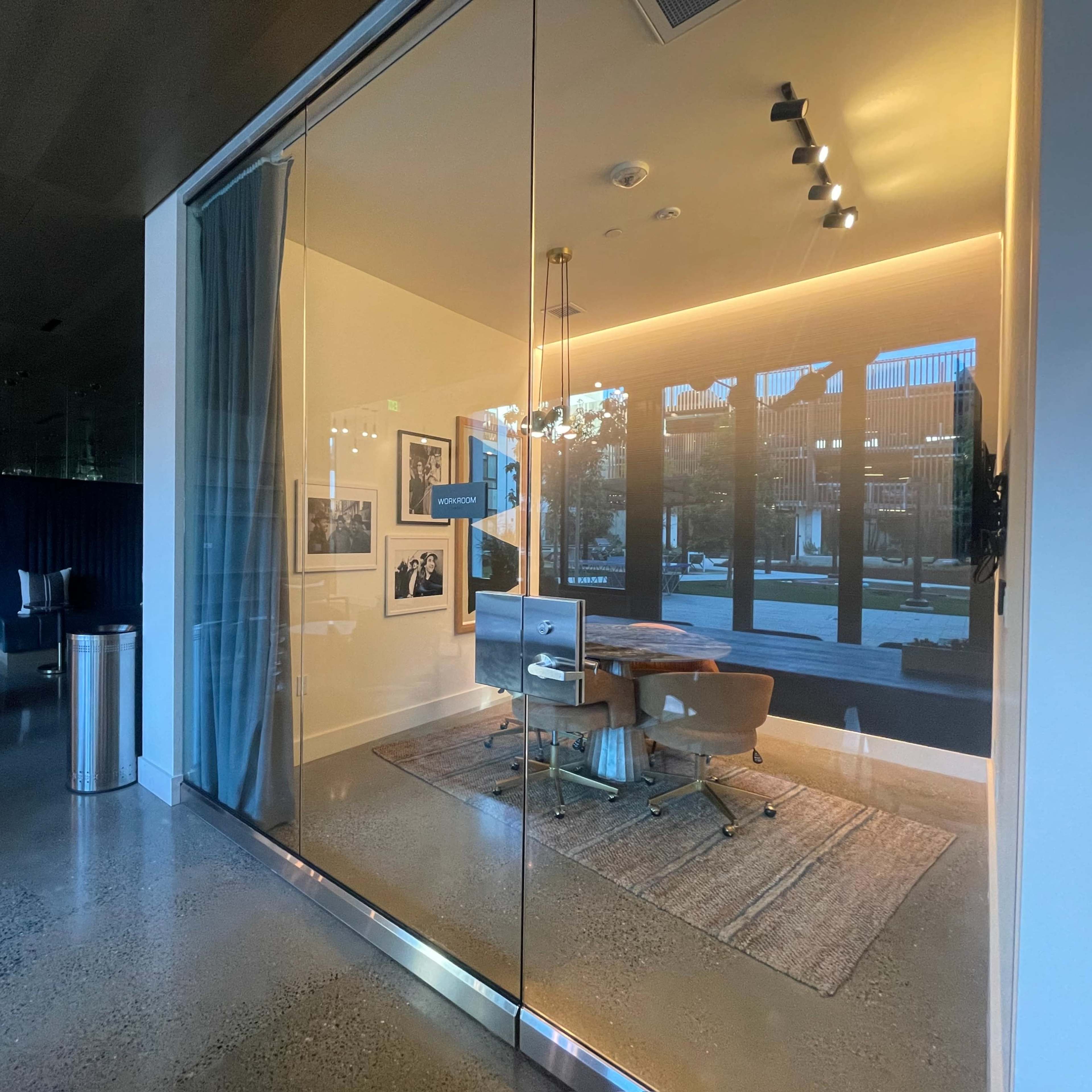 A modern office space with a glass wall, featuring a desk and chair, illuminated by warm lighting, and framed photographs on the wall.