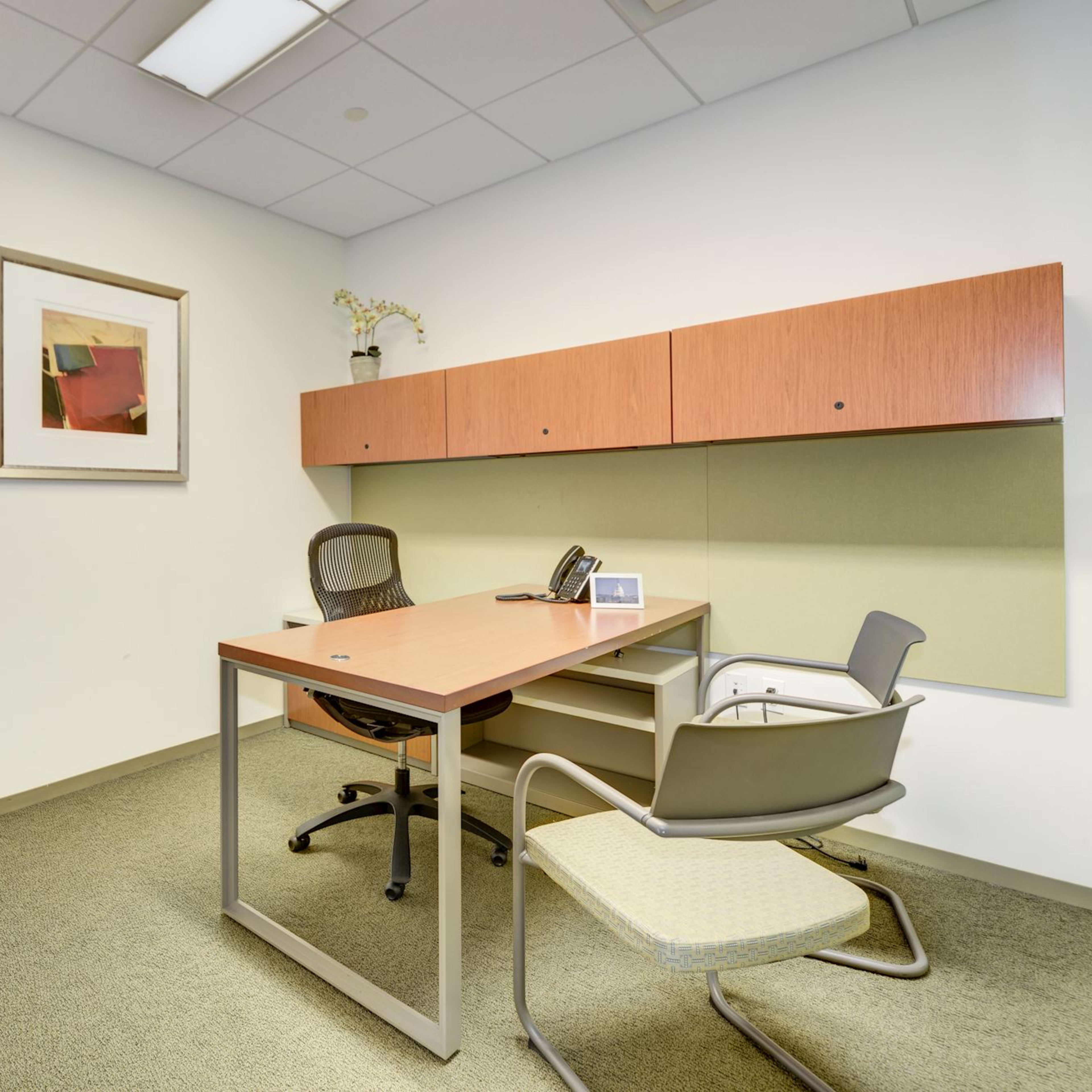 A small office space with a wooden desk, two chairs, a phone, and wall-mounted cabinets.