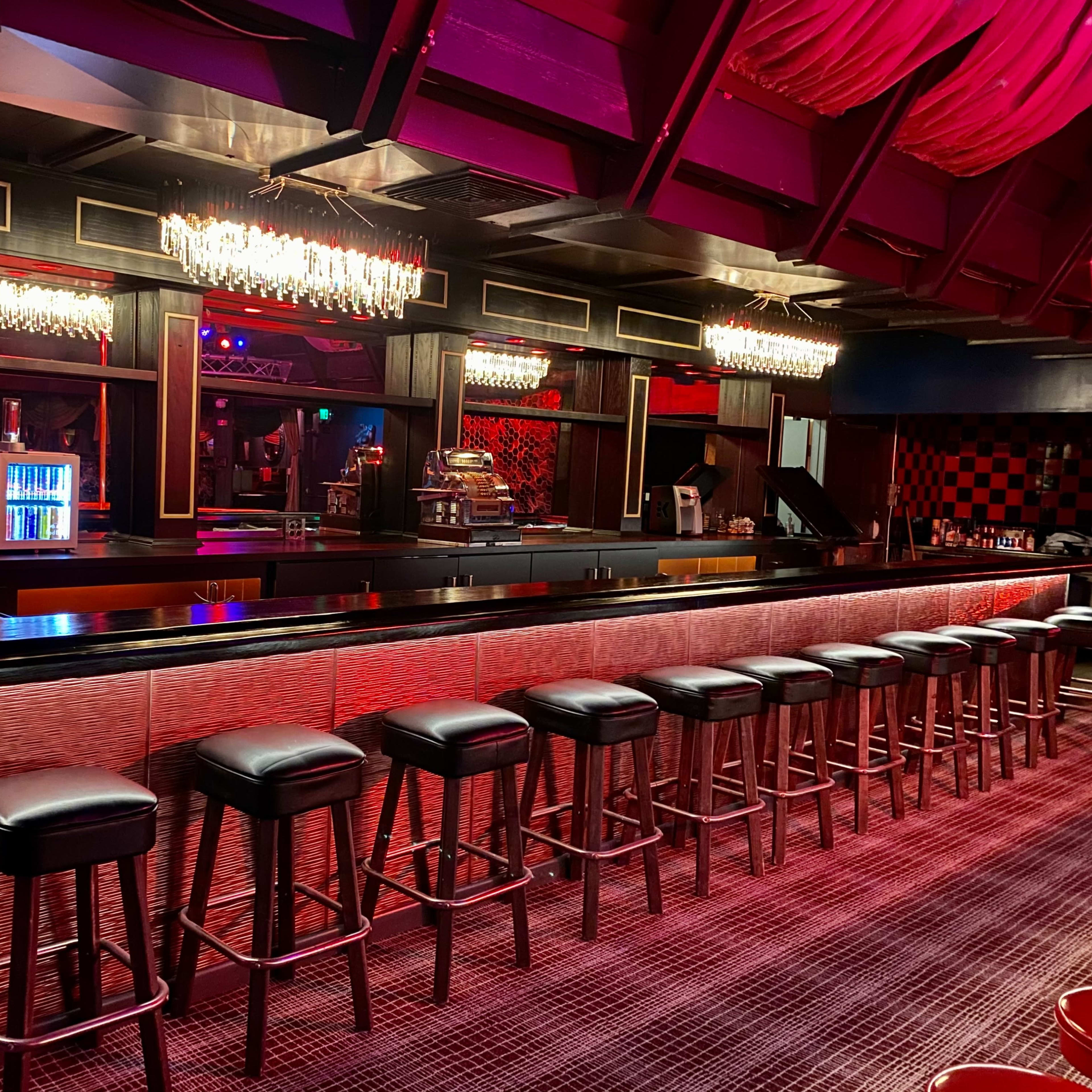 A dimly lit bar with a long counter, rows of bar stools, and crystal chandeliers overhead.