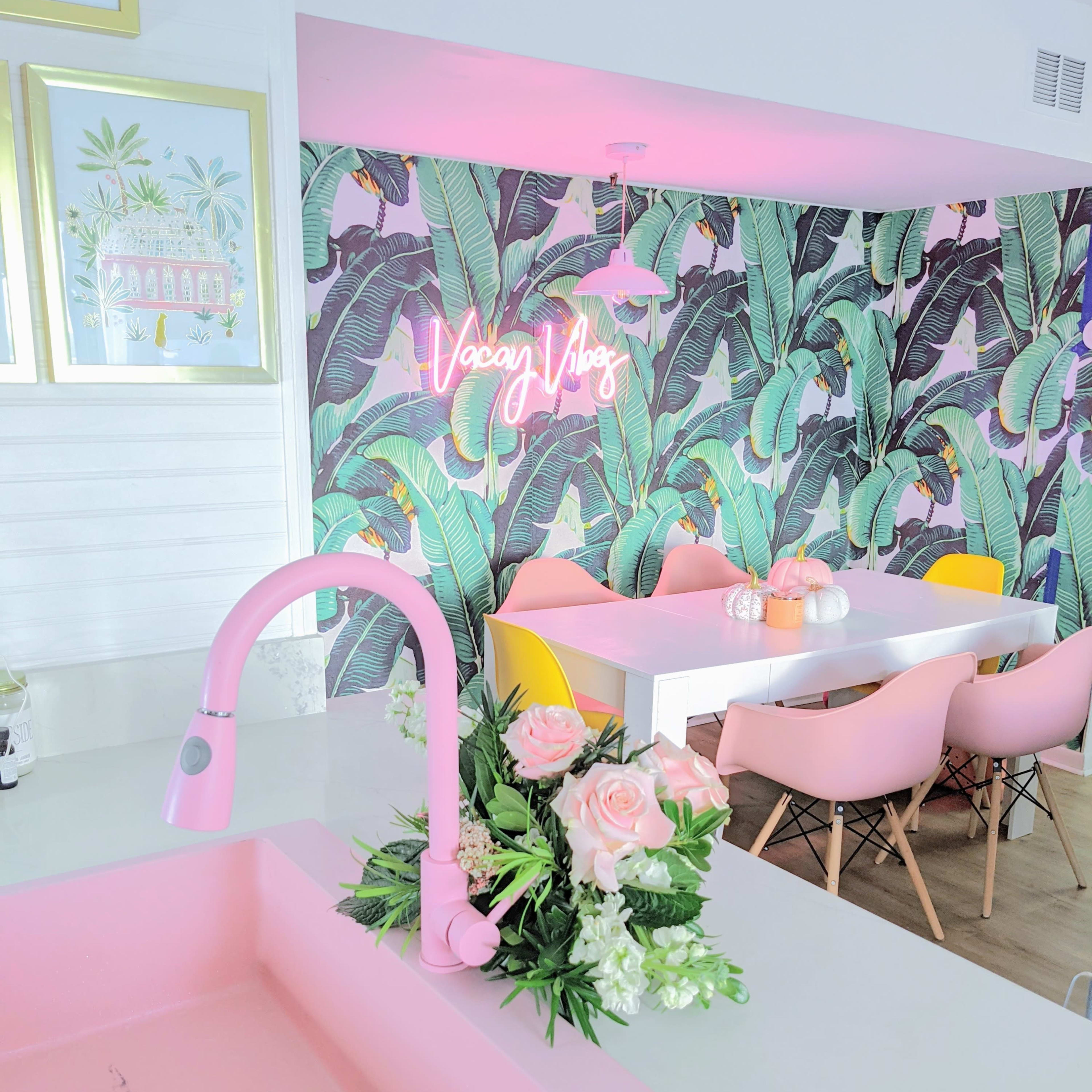 A modern kitchen features a pink sink and faucet, complemented by tropical wallpaper and a dining area with colorful chairs and a neon sign.