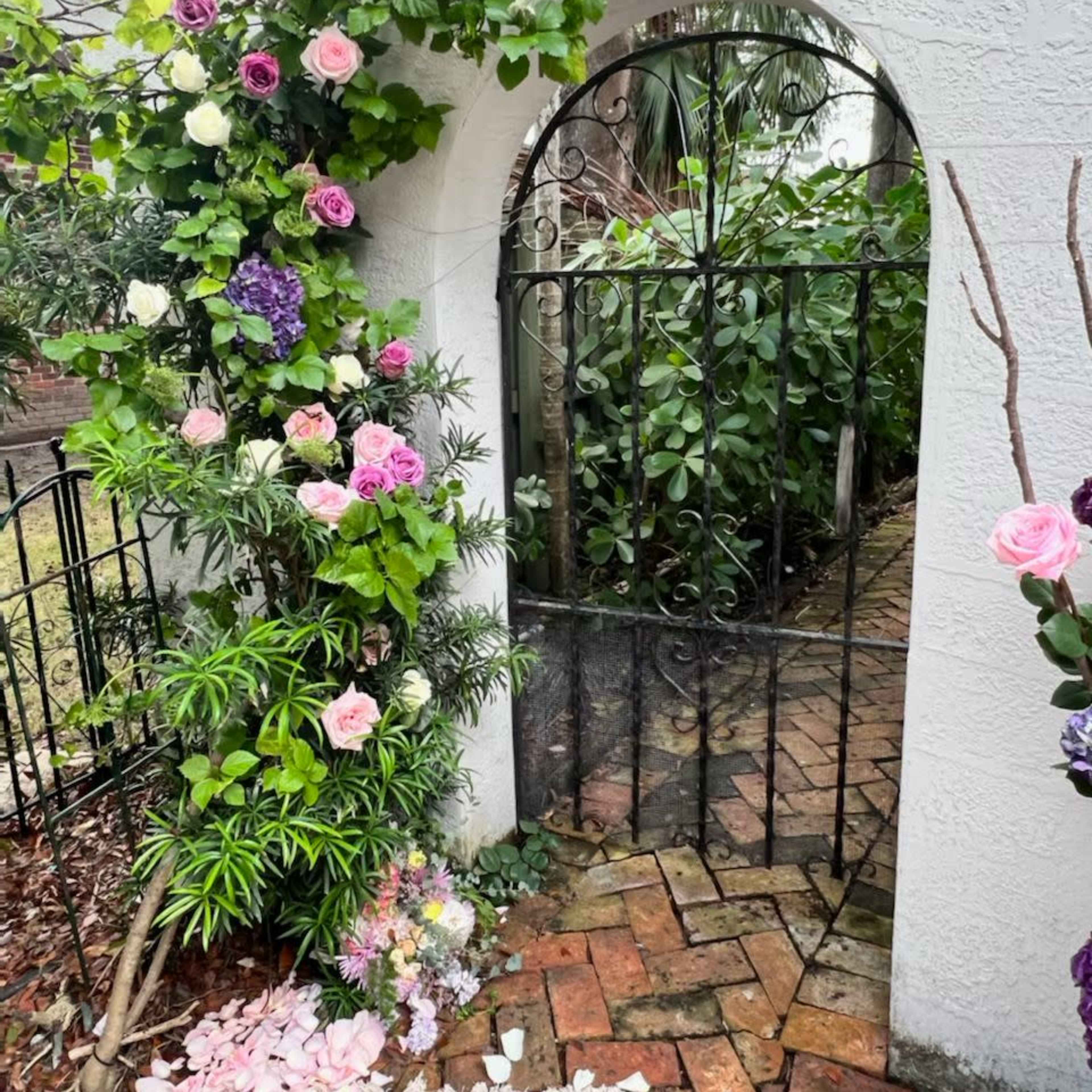 A black wrought iron gate is framed by climbing plants and flowers, opening into a lush garden pathway lined with brick.