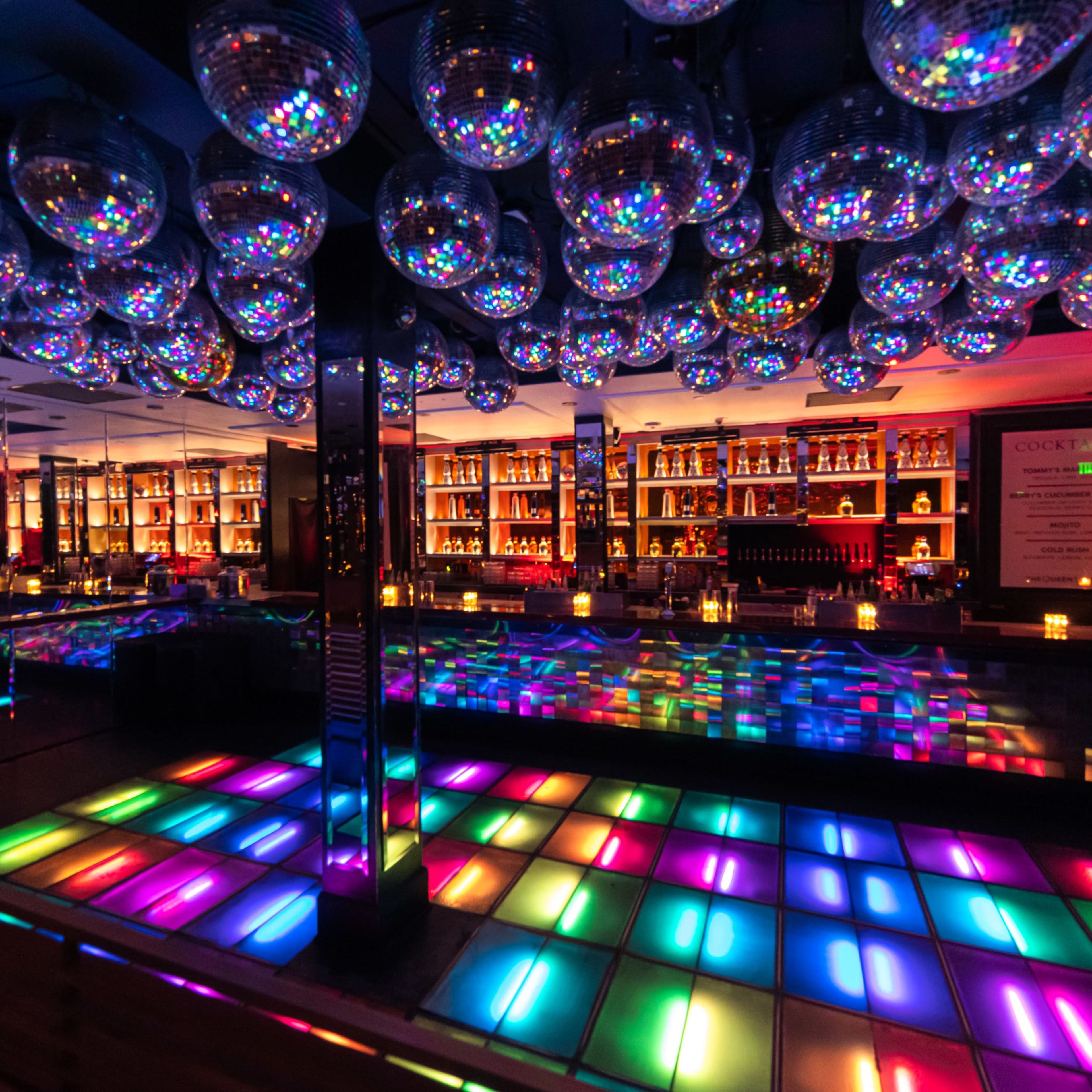 A vibrant dance floor illuminated with colorful lights, surrounded by mirrored disco balls and a well-stocked bar in the background.