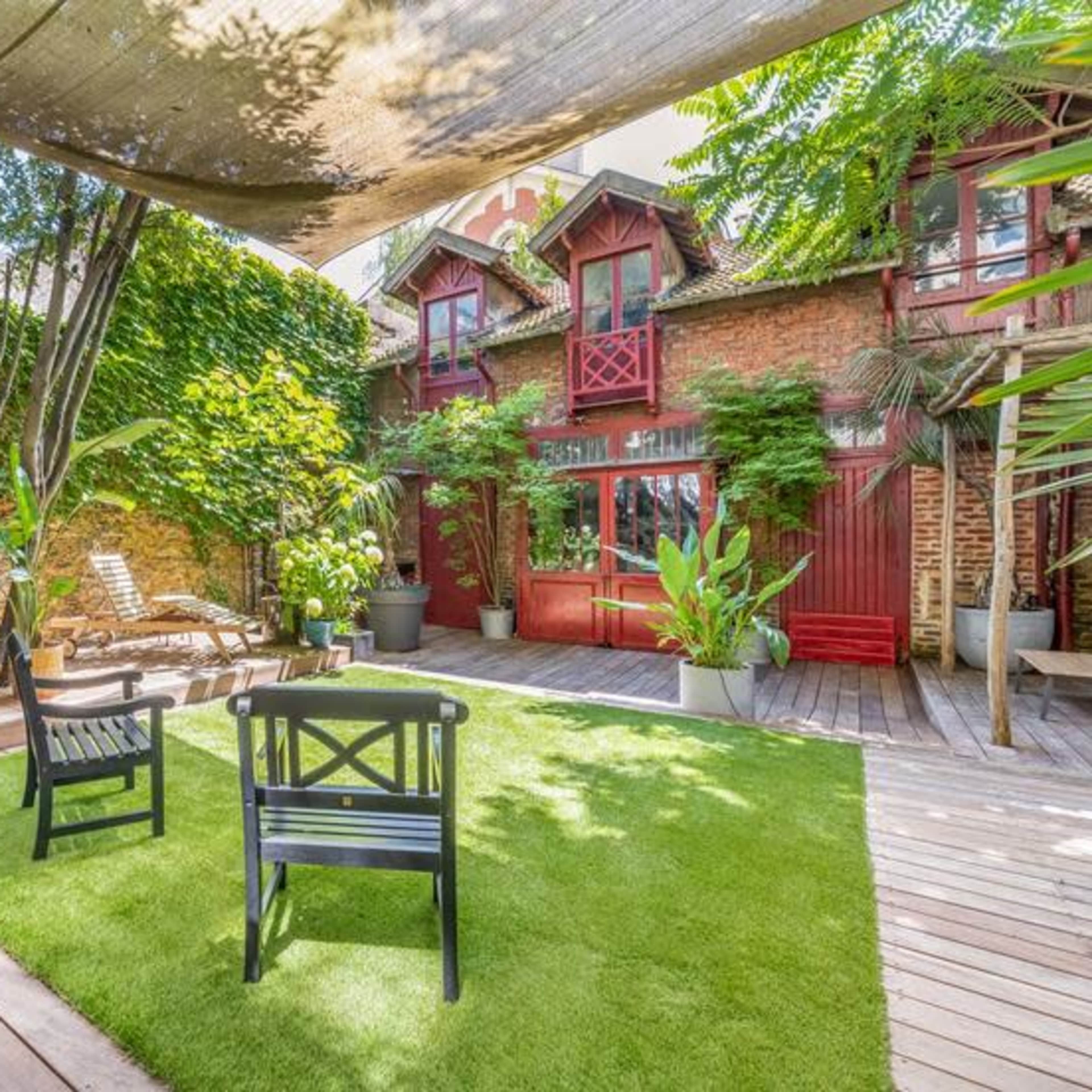 A well-maintained garden area with wooden decking, green grass, and two black chairs surrounded by vibrant plants and a red-brick building in the background.
