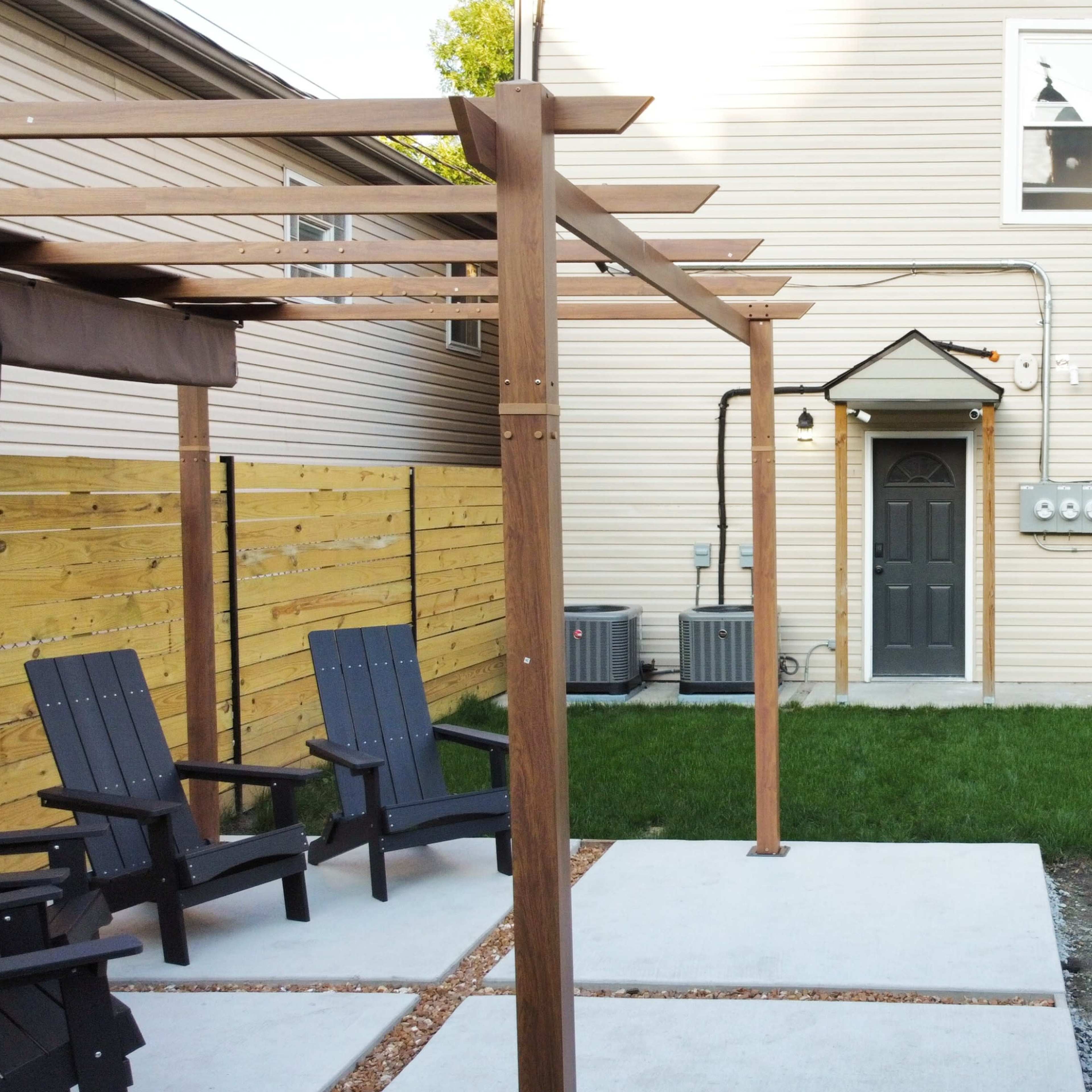 A backyard with a pergola, two black chairs, a barbecue grill, and a neatly maintained patch of grass adjacent to a house.