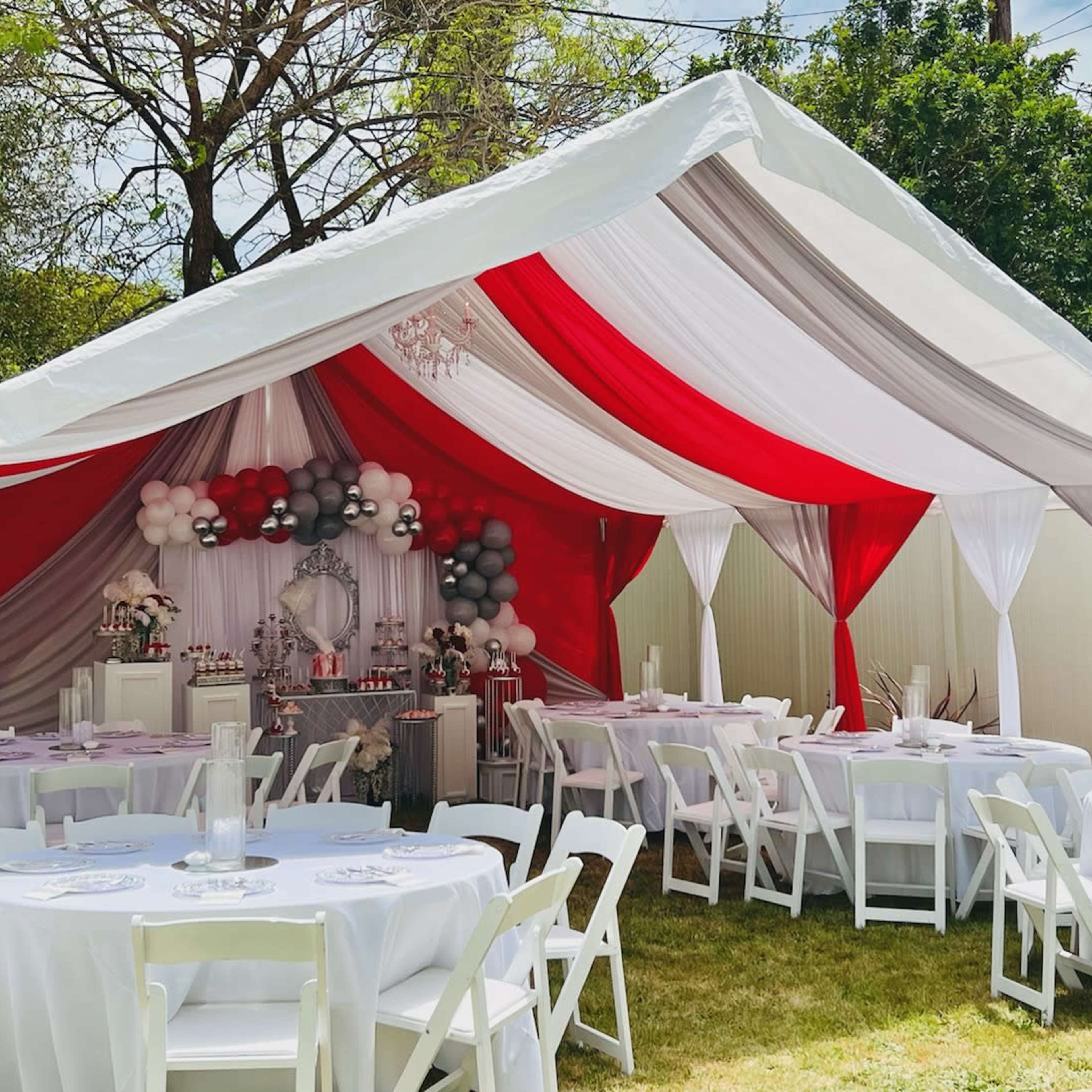 A decorated outdoor tent with red, white, and gray drapes houses tables set with white linens and contains festive decor, including balloons and a dessert display.
