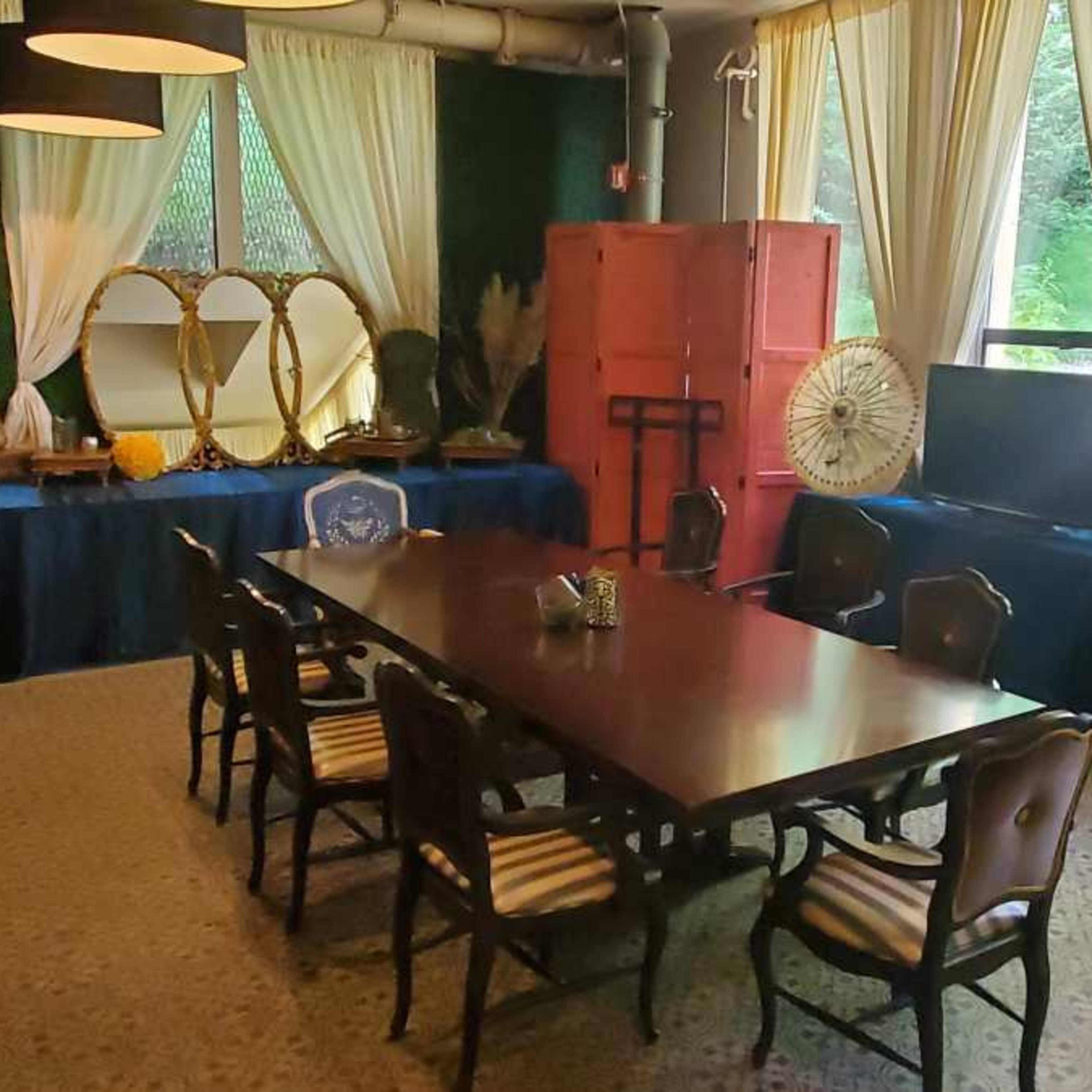 A room furnished with a large wooden table surrounded by chairs, two televisions on a blue-covered surface, and decorative elements including plants and curtains.
