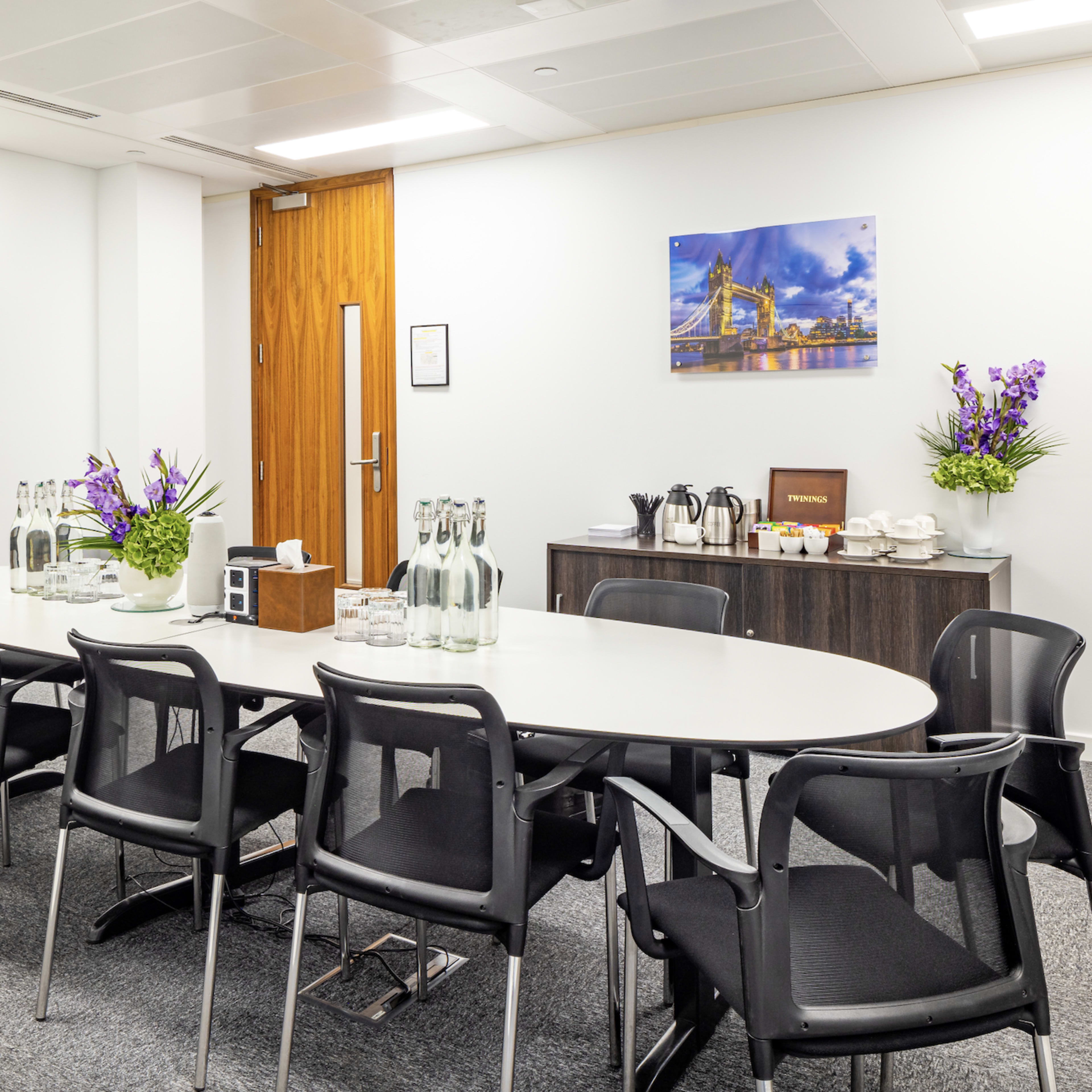 A modern conference room features a long oval table surrounded by black chairs, with a television mounted on the wall and refreshments set on a side cabinet.