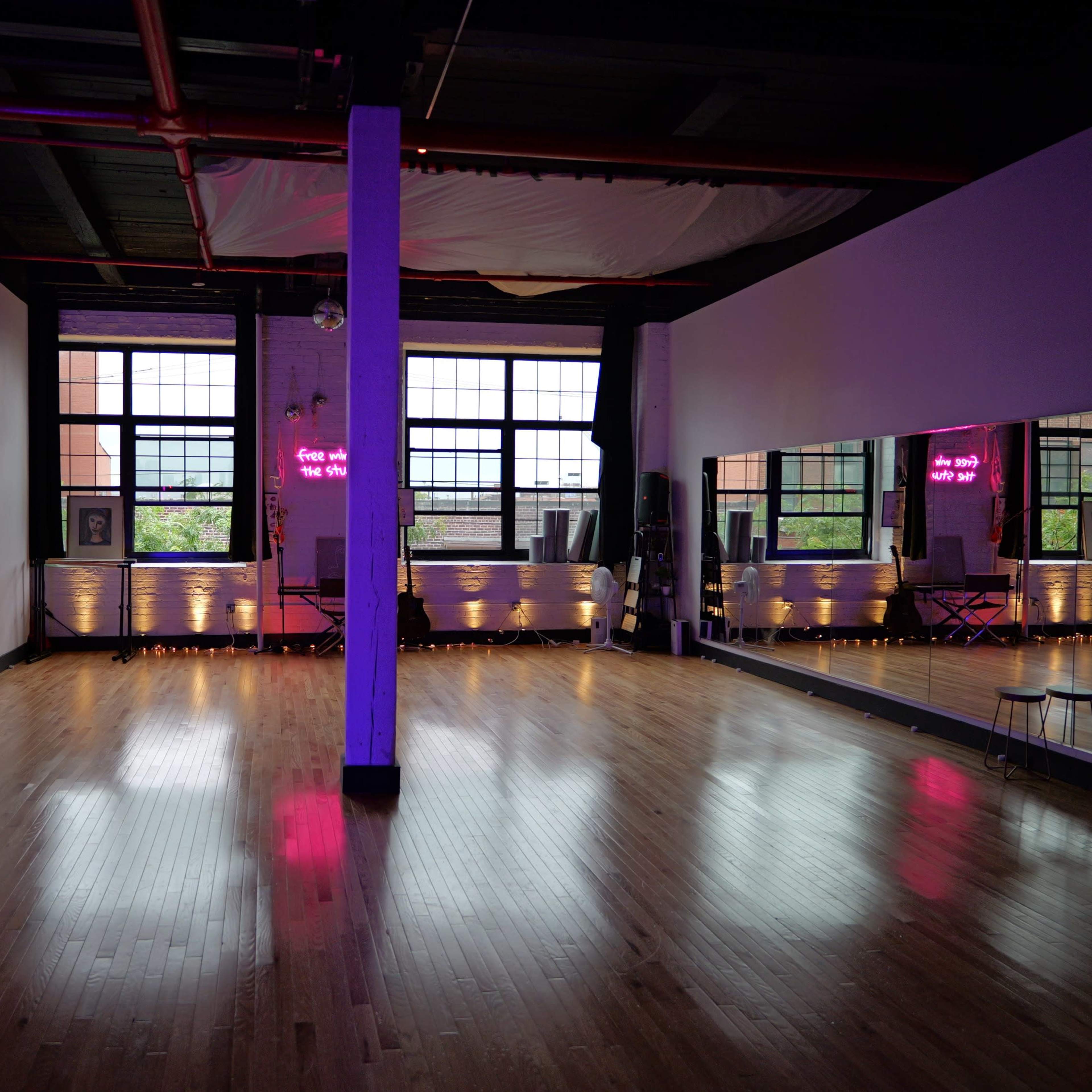 A spacious dance studio with hardwood floors, large windows, and neon signs illuminating the walls.