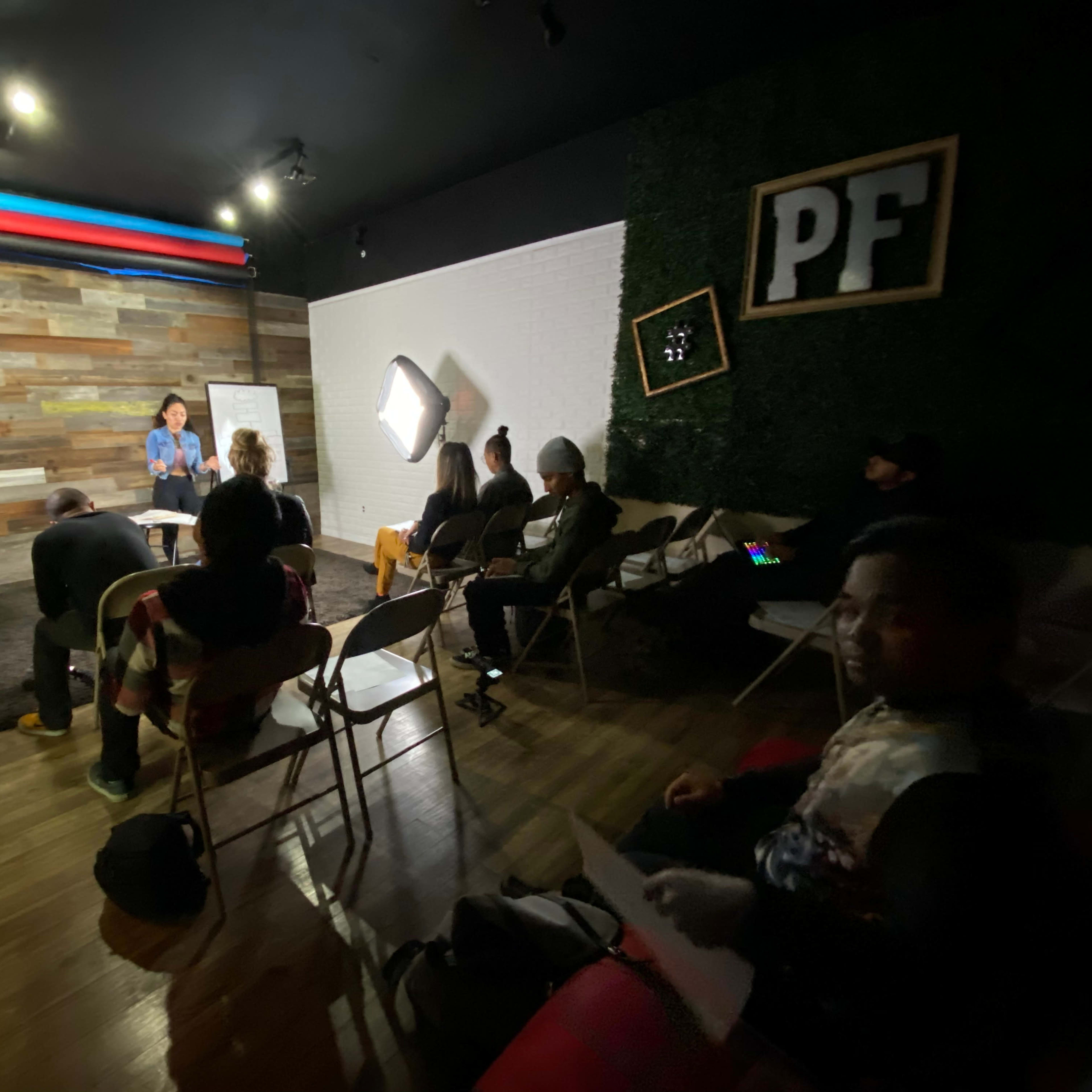 A group of people is seated in a room with wooden walls, listening to a presentation from a woman standing in front of a whiteboard.