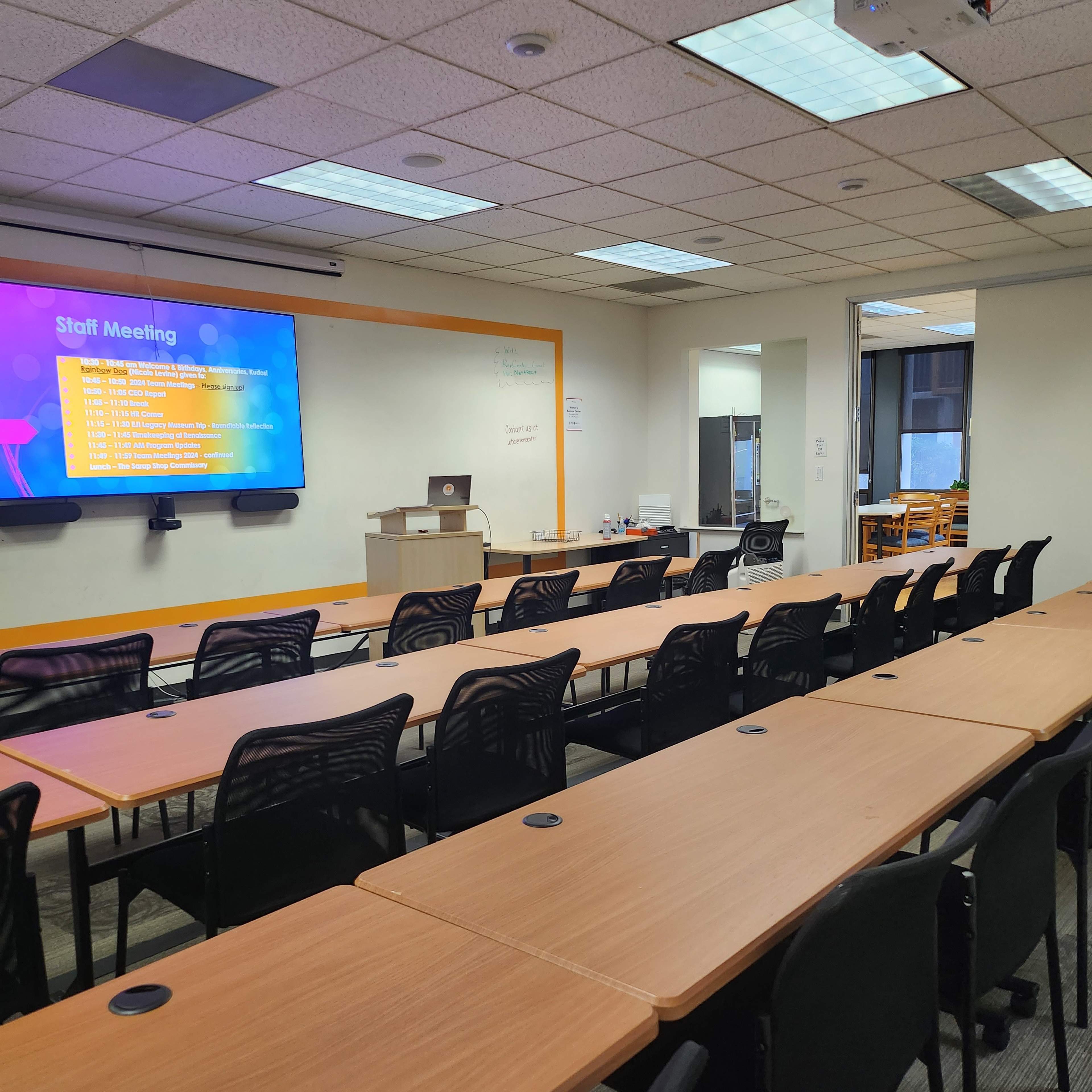 A conference room is set up with rows of tables and chairs facing a projector screen displaying a meeting agenda.