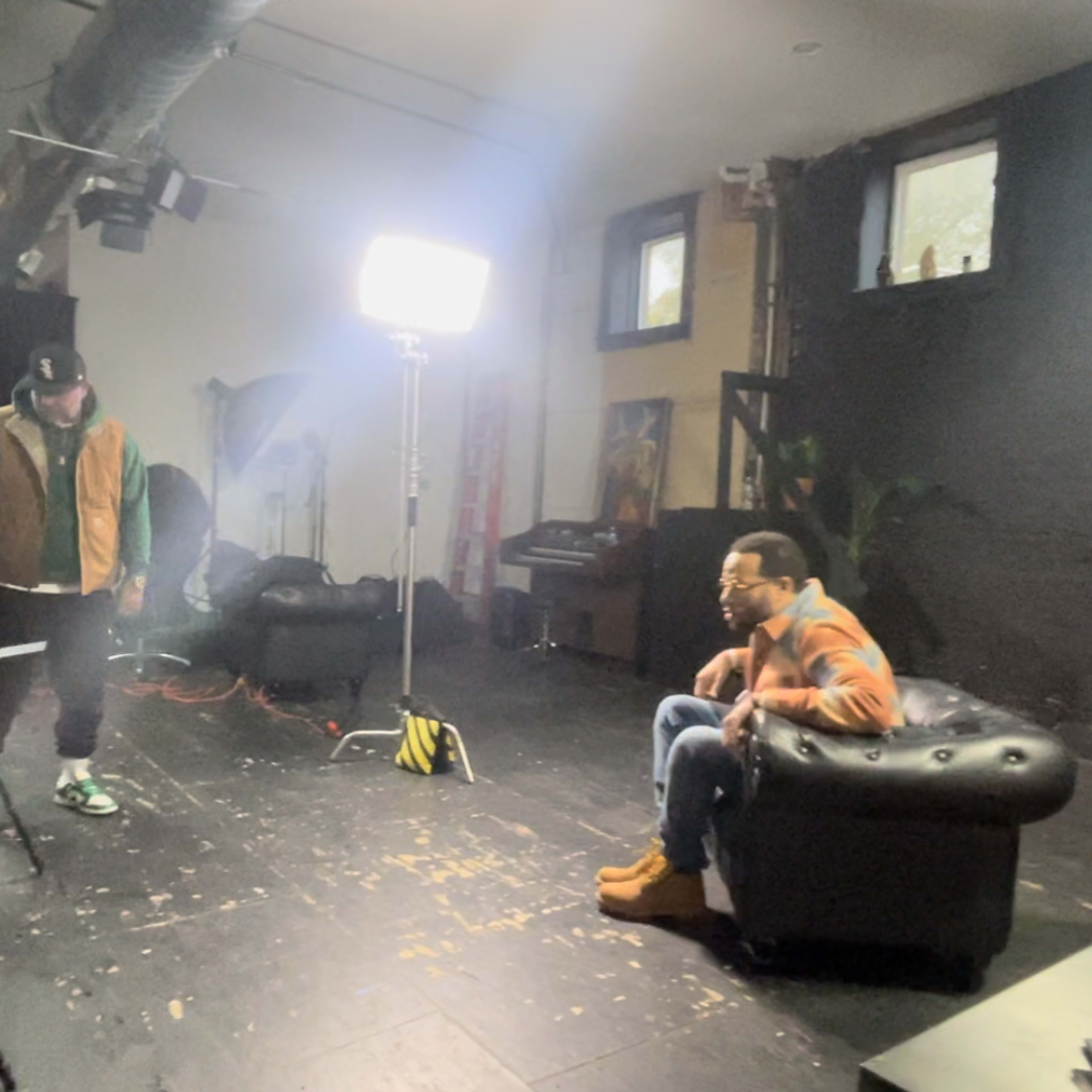A group of three people is in a dimly lit studio setting with lighting equipment, cameras, and a black couch.