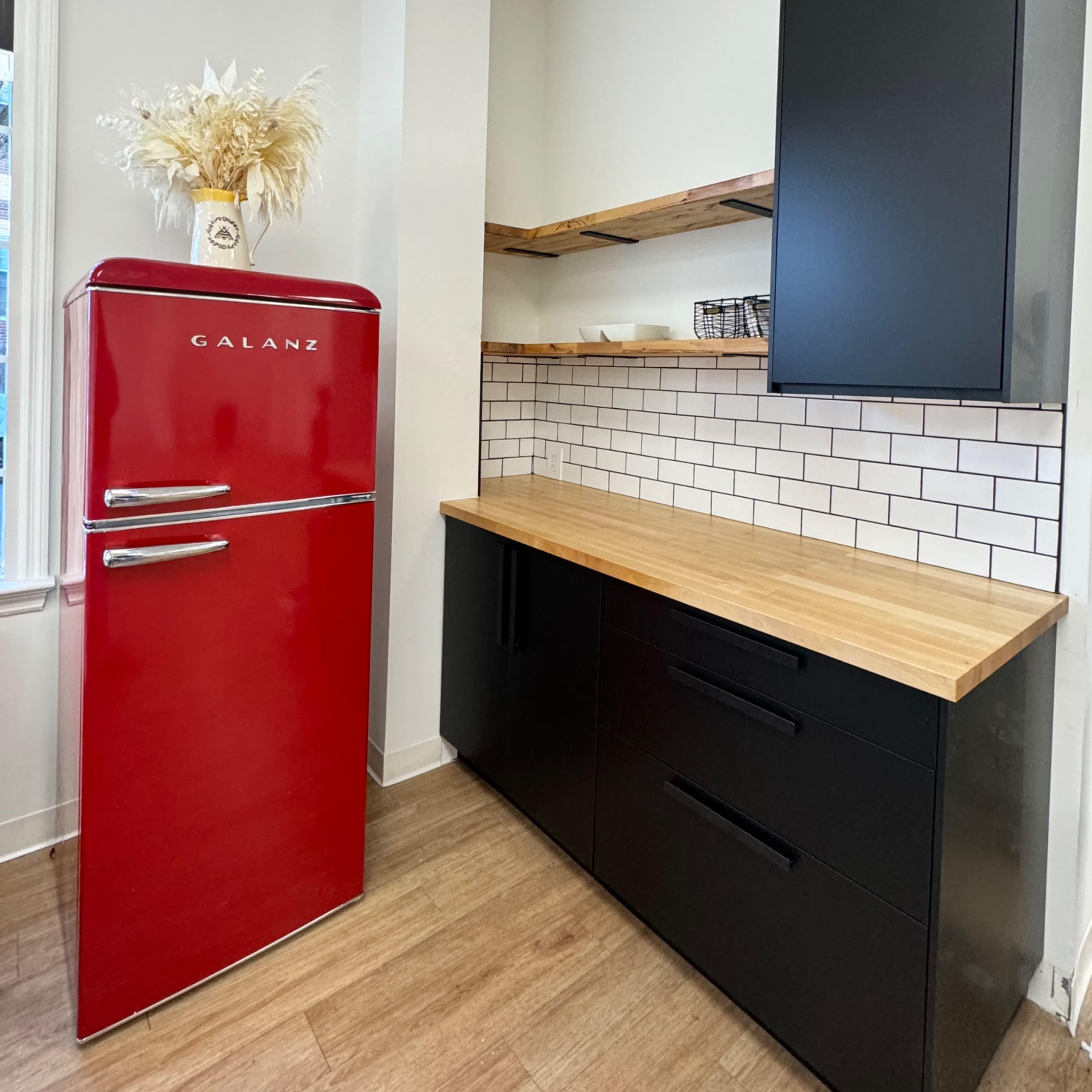 A modern kitchen featuring a red refrigerator, wooden countertops, and black cabinetry against a white tiled backsplash.
