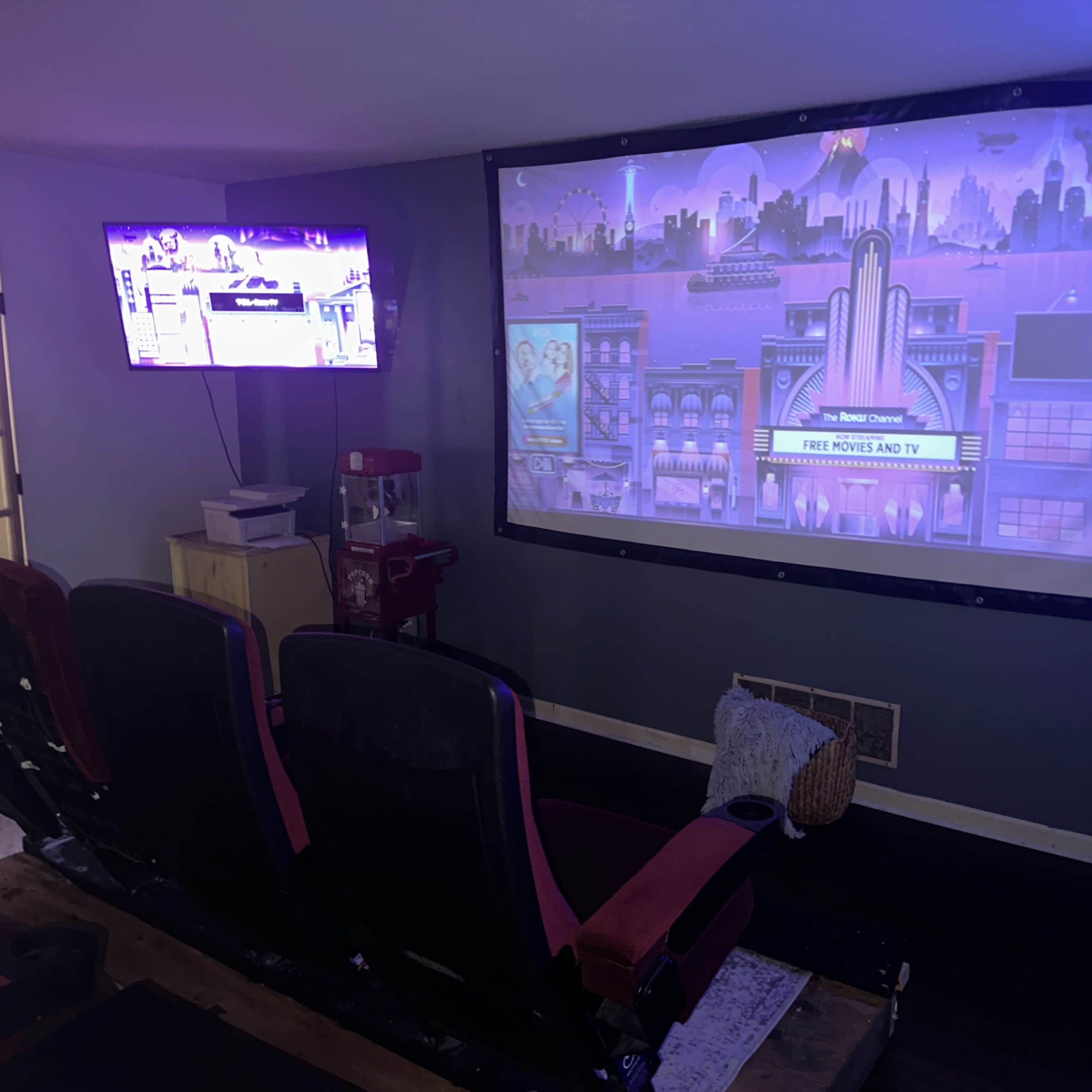 A home theater setup features a large projection screen with a colorful image, a smaller TV on the wall, and red theater-style seating.