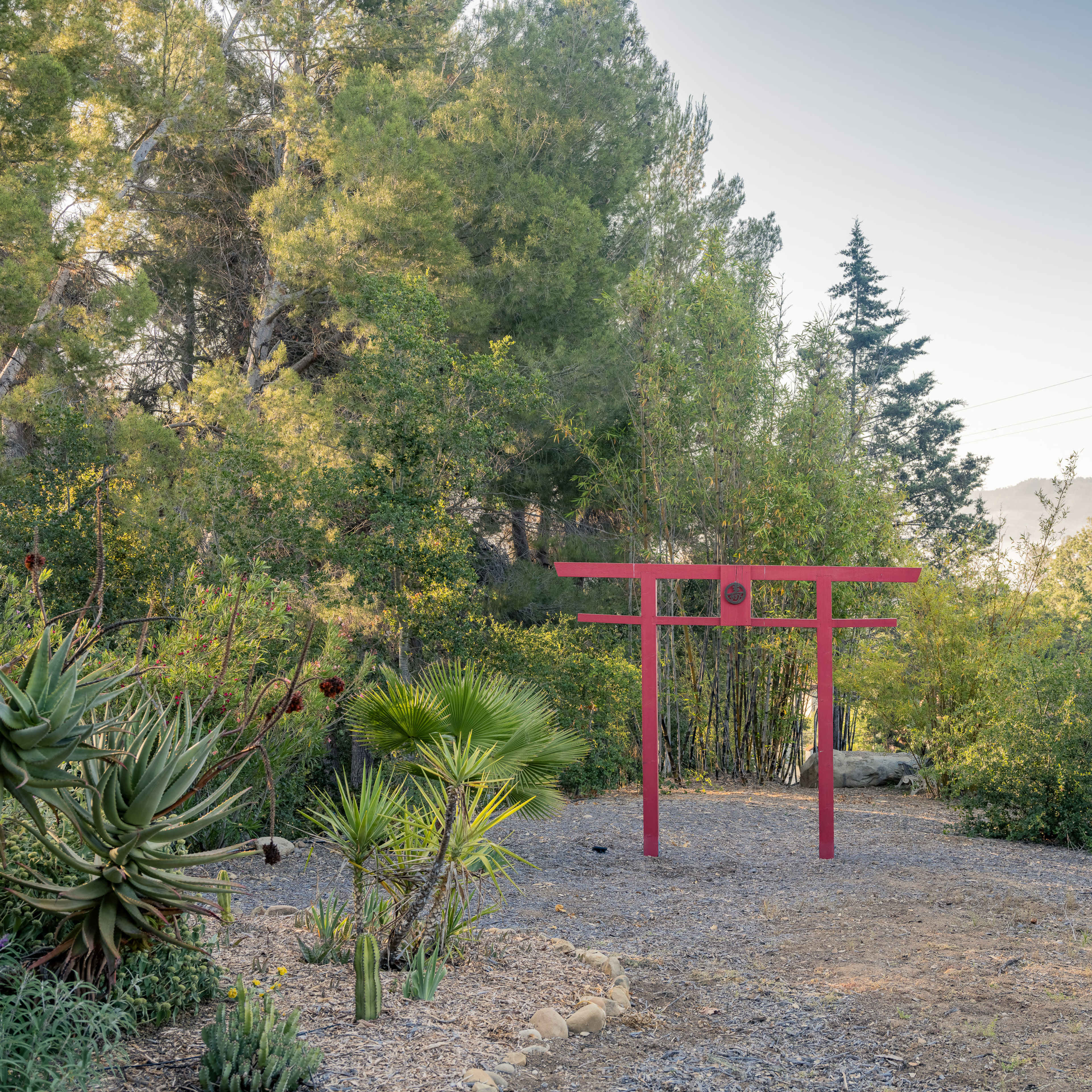 A red torii gate stands in a landscaped area surrounded by various trees and plants.