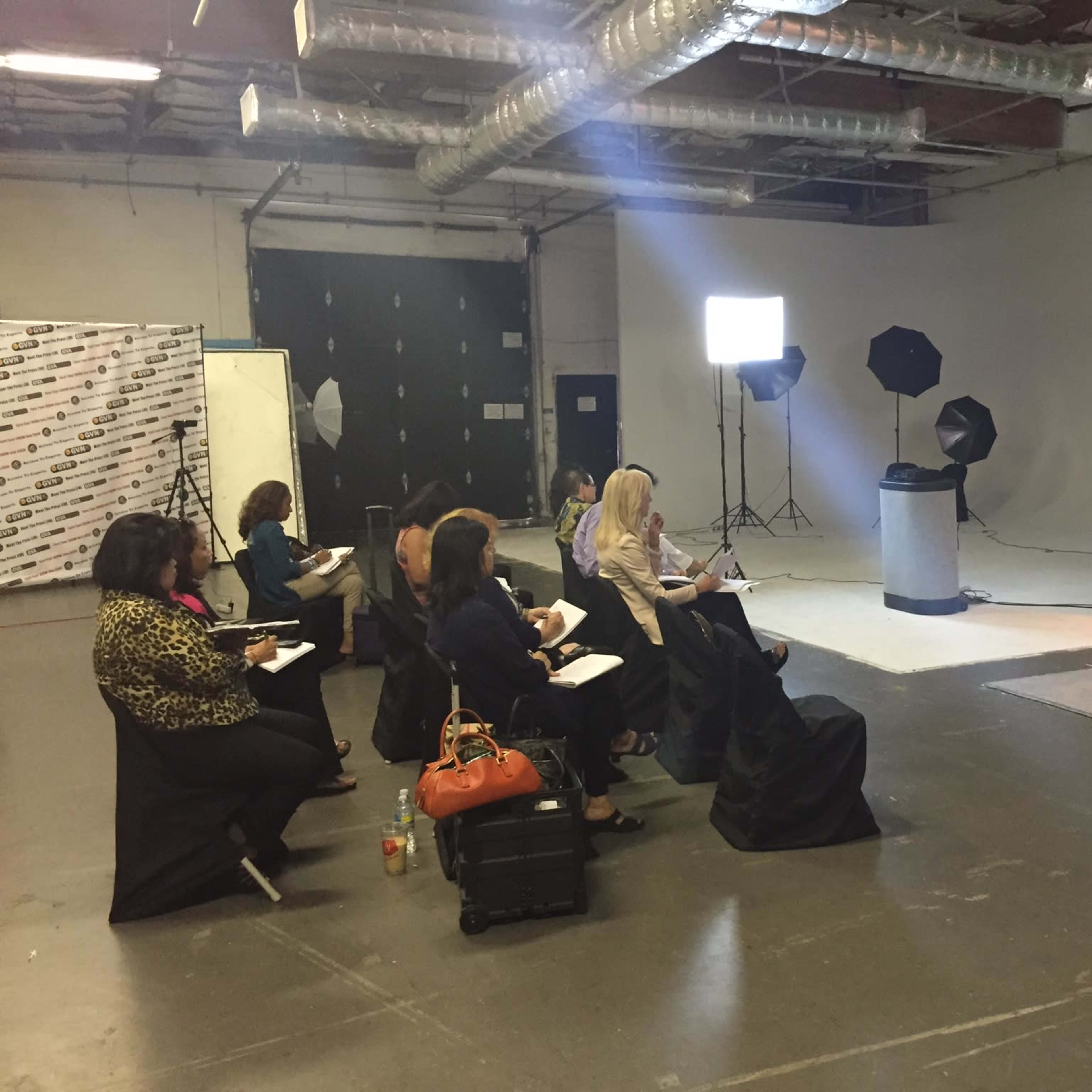 A presenter stands in front of a seated audience in a studio setting, delivering a lecture while attendees take notes.