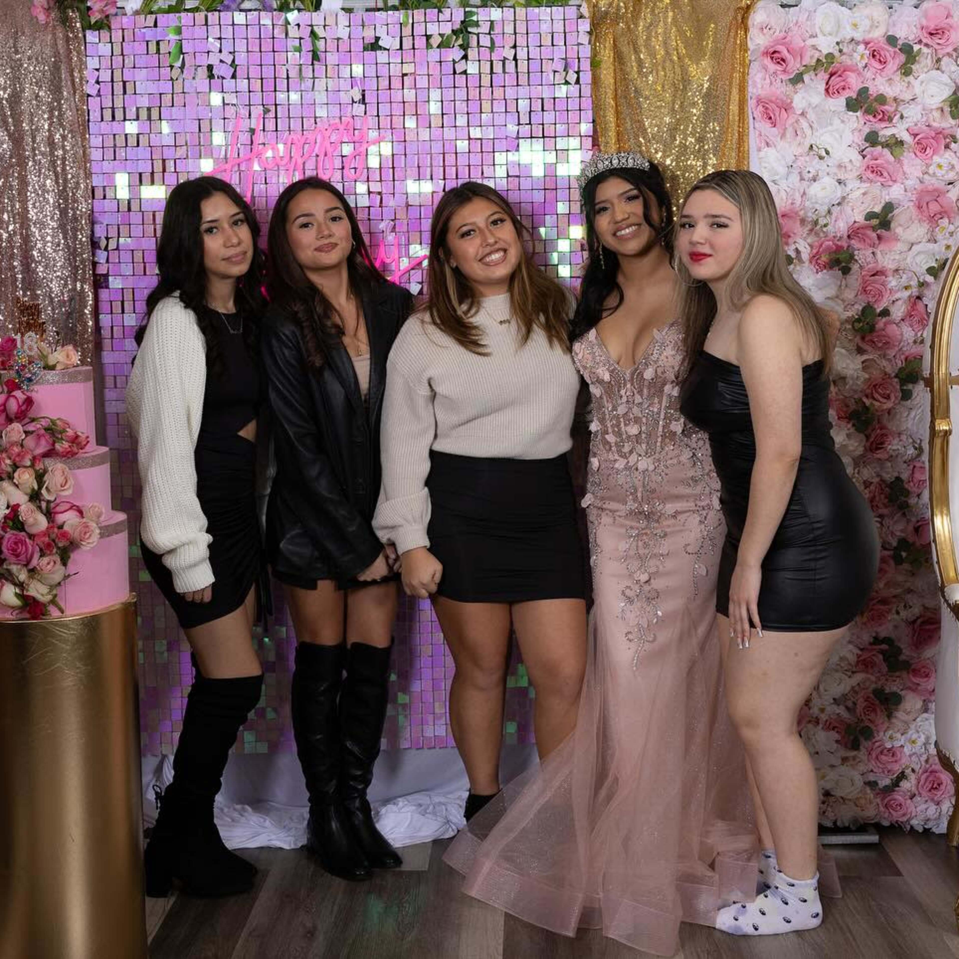 A group of five women poses together in a decorated party area featuring balloons, a floral backdrop, and a cake display.