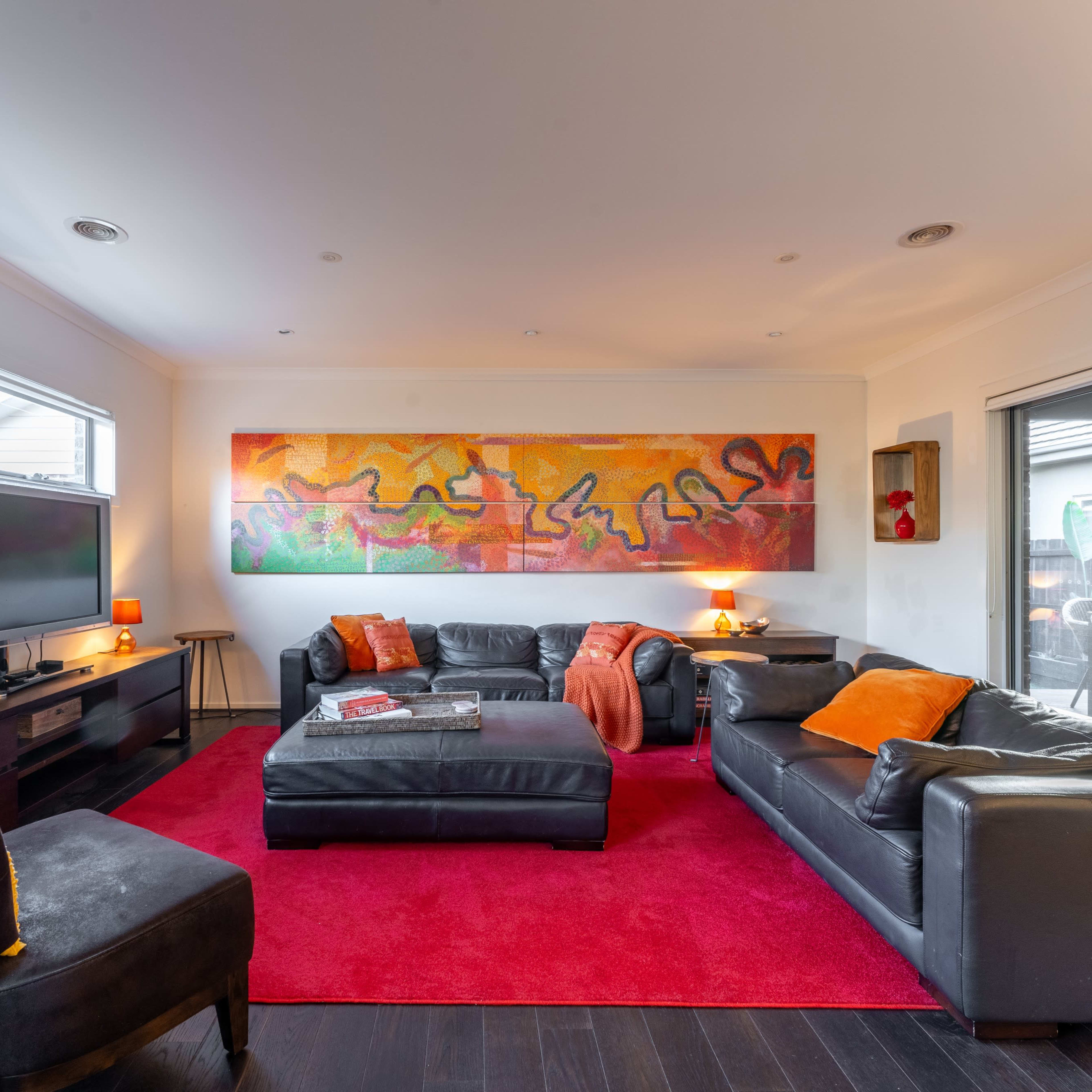 A living room features dark leather furniture, a large colorful artwork on the wall, and a red area rug.