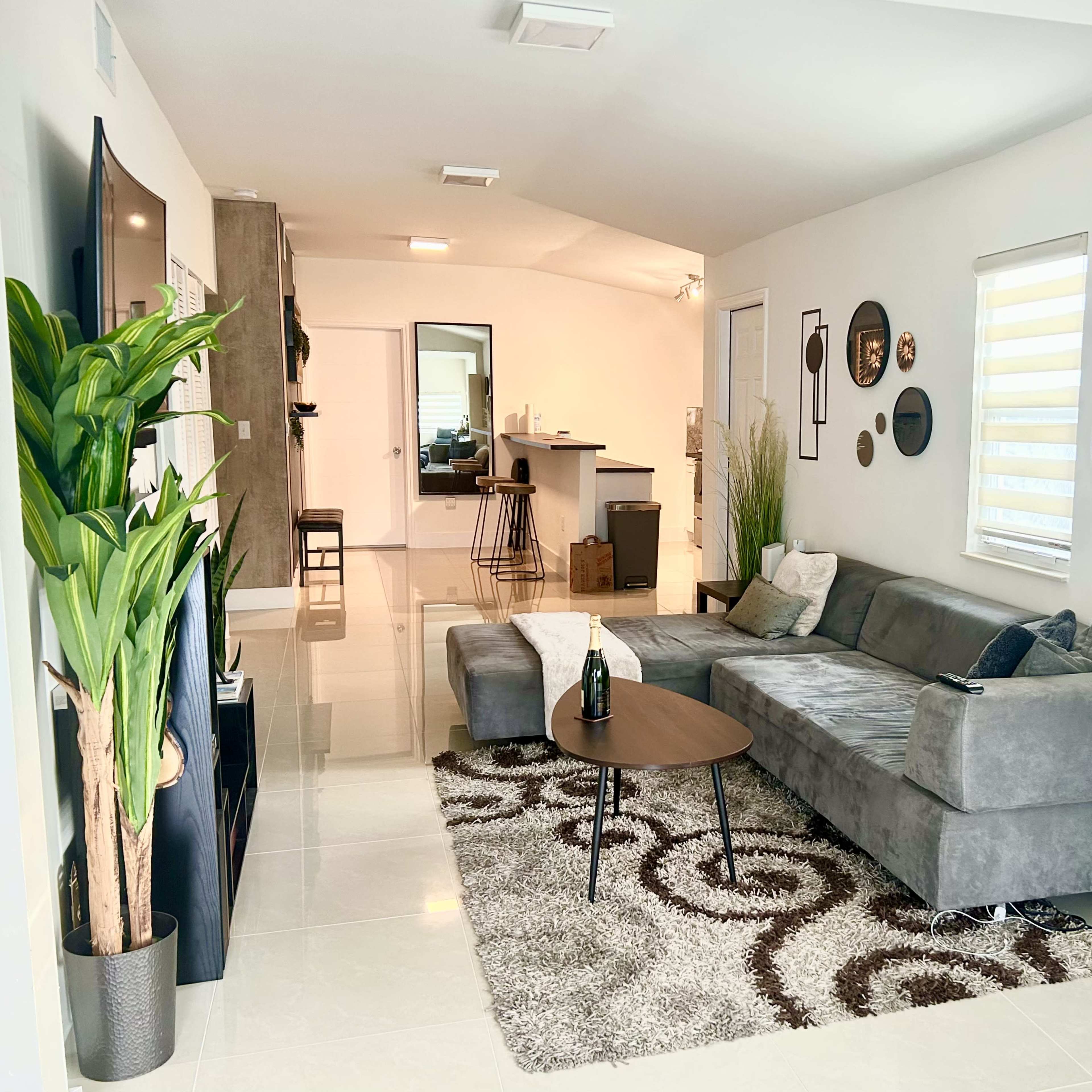 The living room features a gray sectional sofa, a round coffee table, and a decorative rug, with a kitchen area visible in the background.