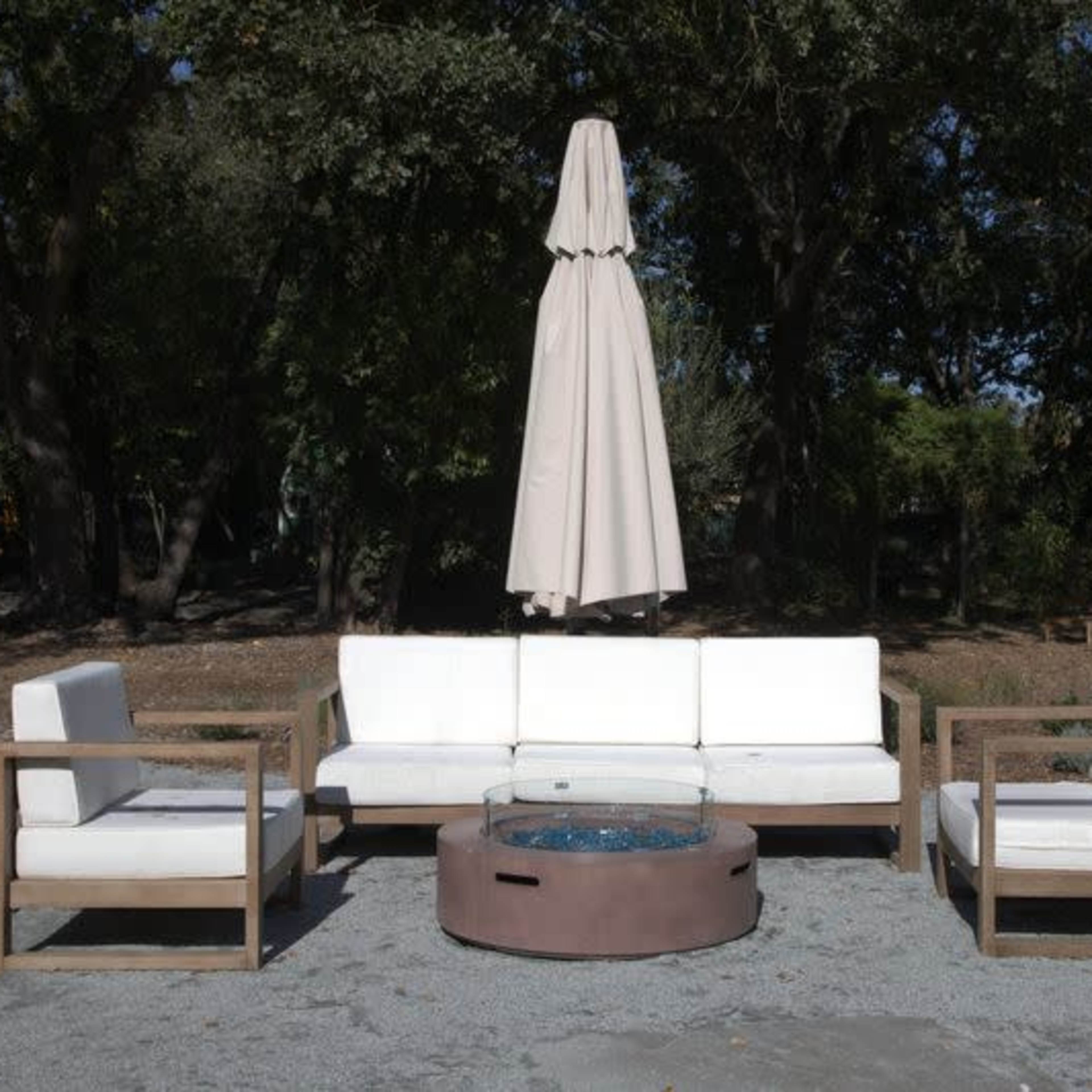 A modern outdoor seating arrangement features a sofa and two armchairs surrounding a circular fire pit, with a large umbrella positioned above.