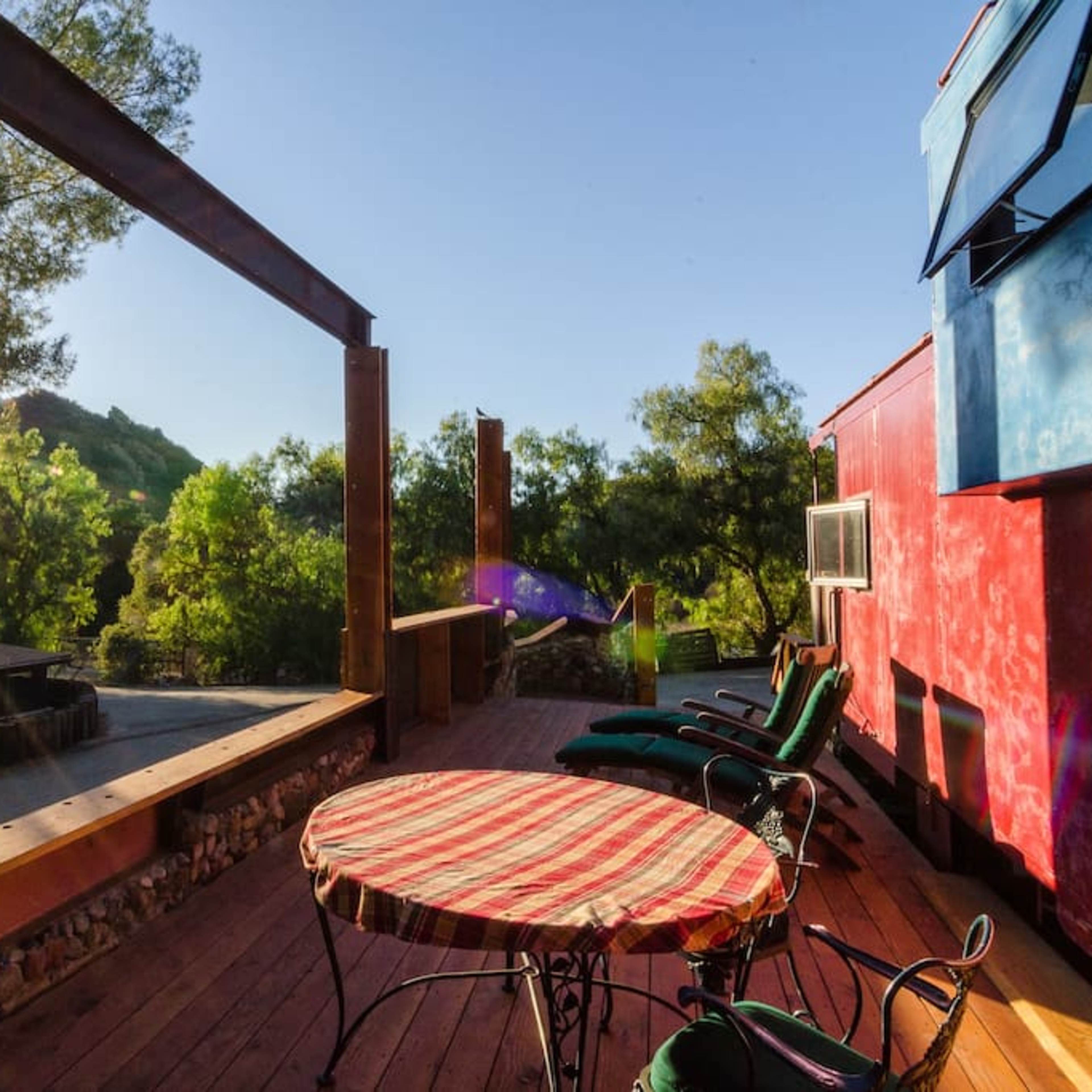 A deck with a round table and lounge chairs overlooks a landscape with trees and mountains, illuminated by sunlight.