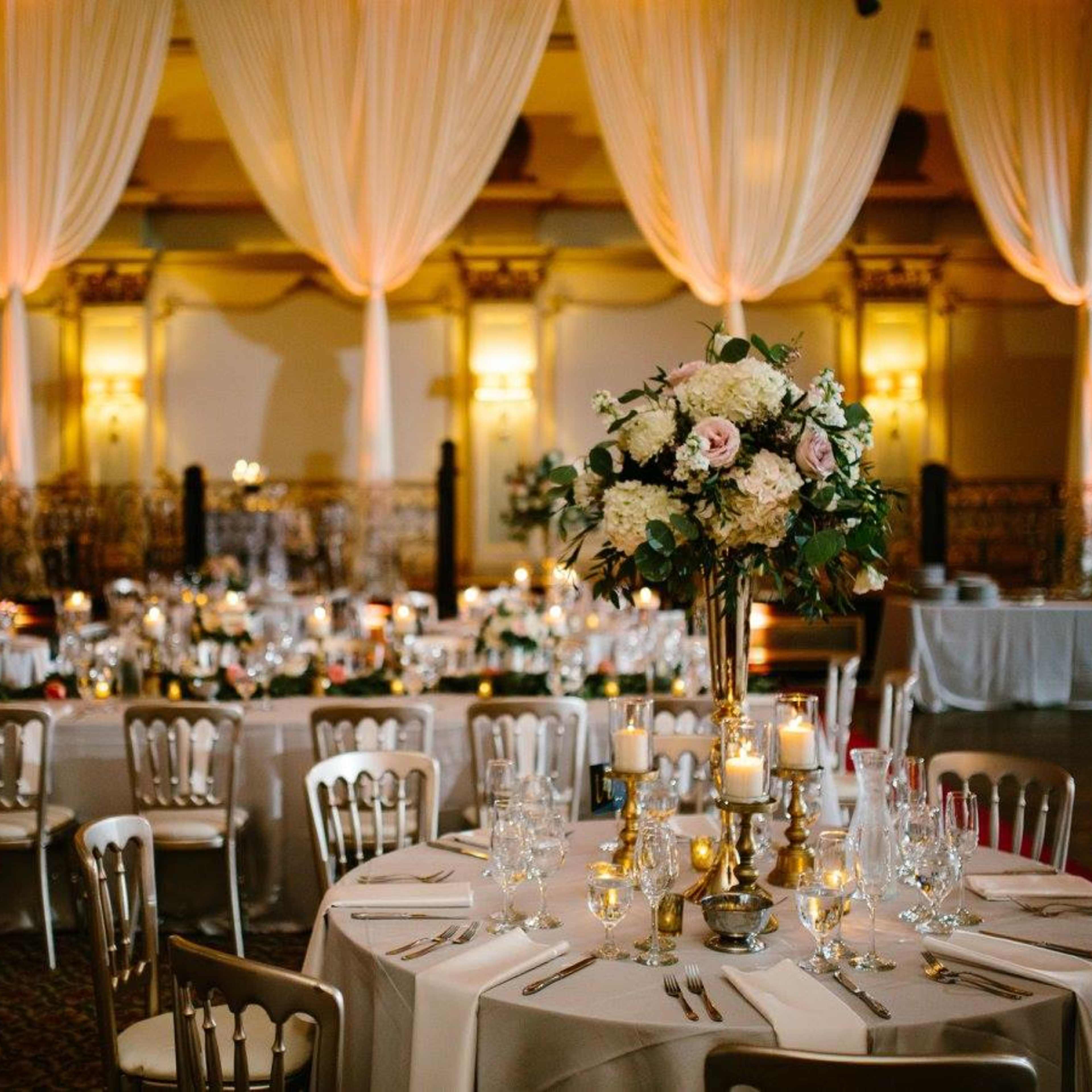 A banquet hall is set for an event, featuring elegantly arranged tables with floral centerpieces, candles, and draped fabric overhead.