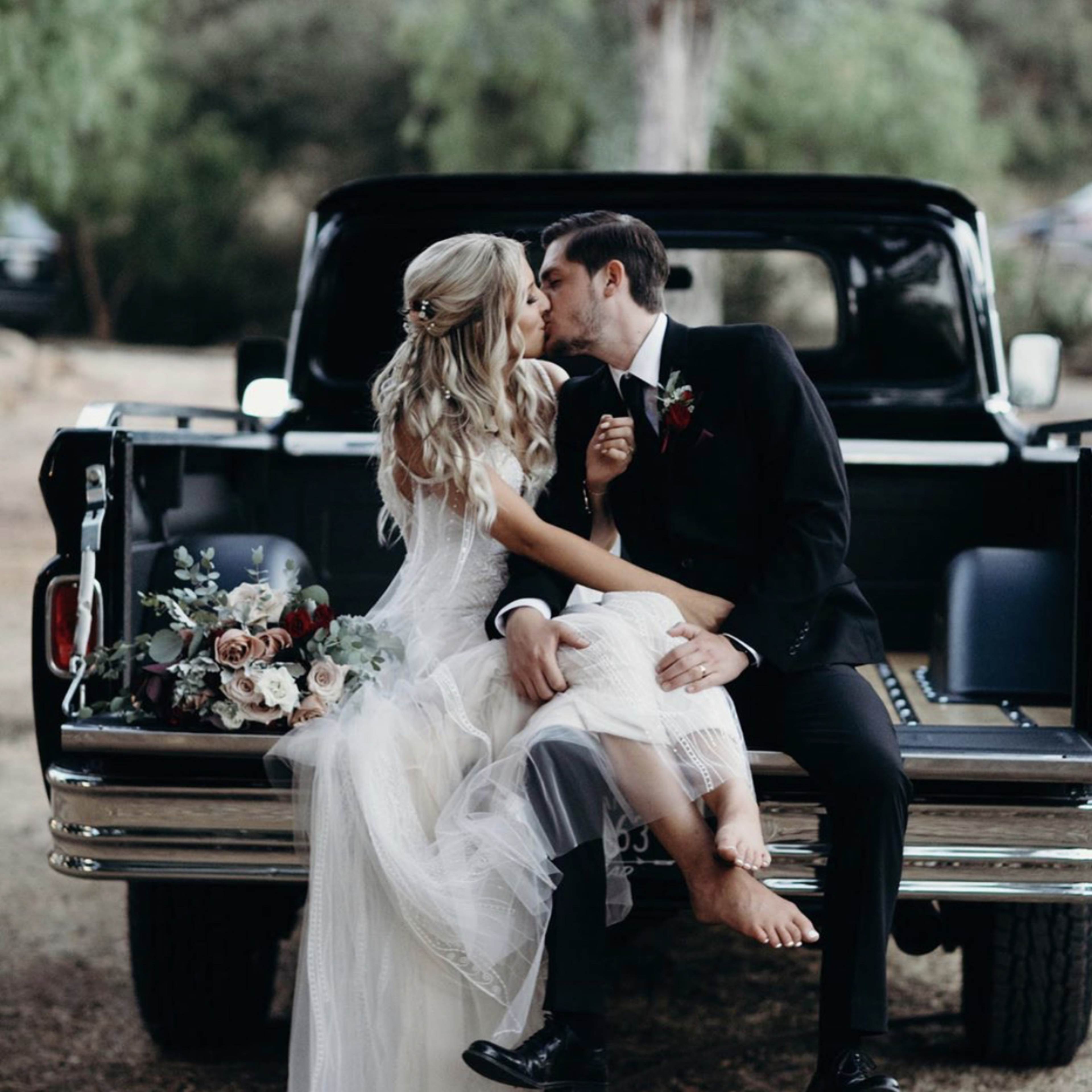 A bride and groom share a kiss while sitting on the back of an old pickup truck, surrounded by greenery.