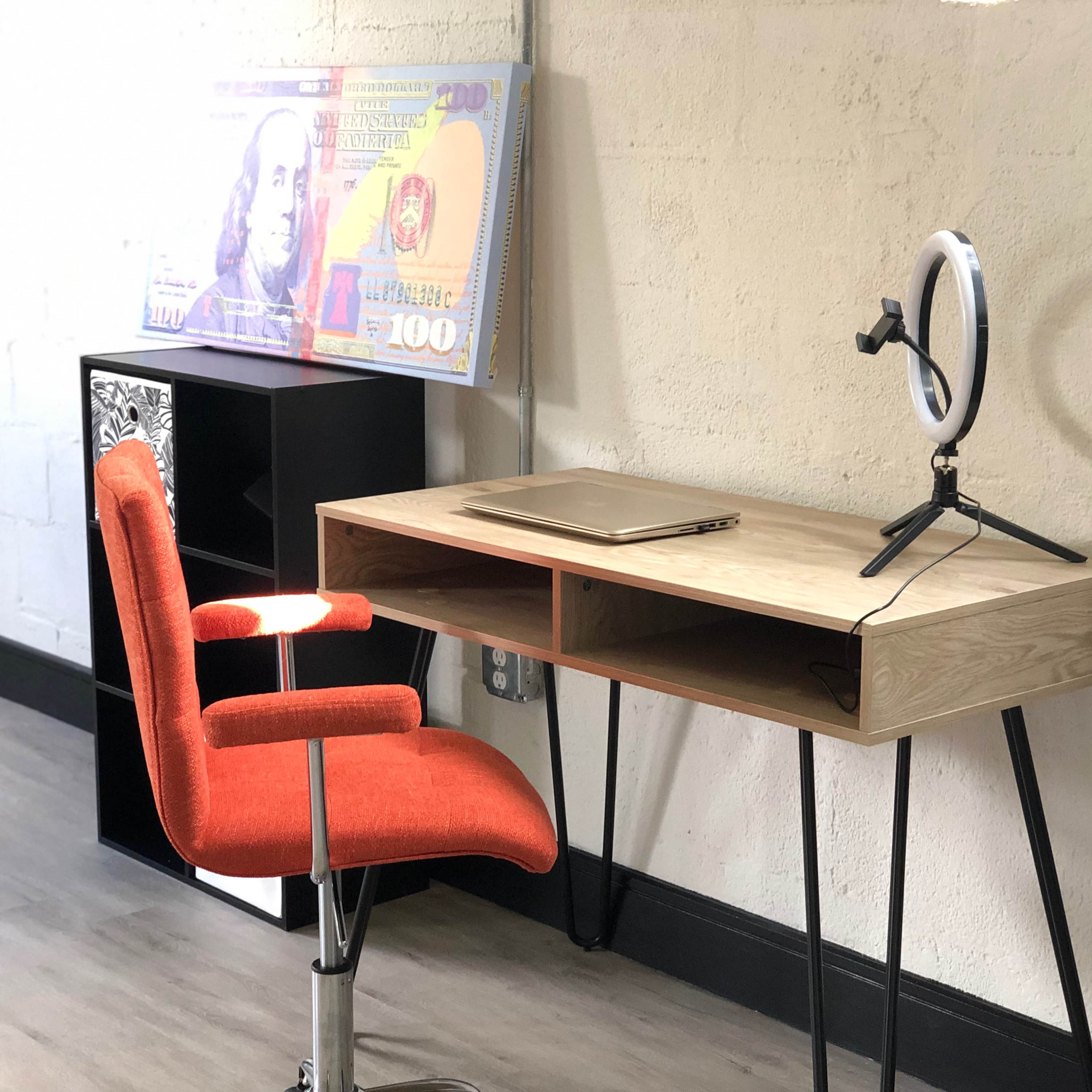 A modern wooden desk with an orange chair and a ring light stands against a white wall, alongside a large decorative banknote.