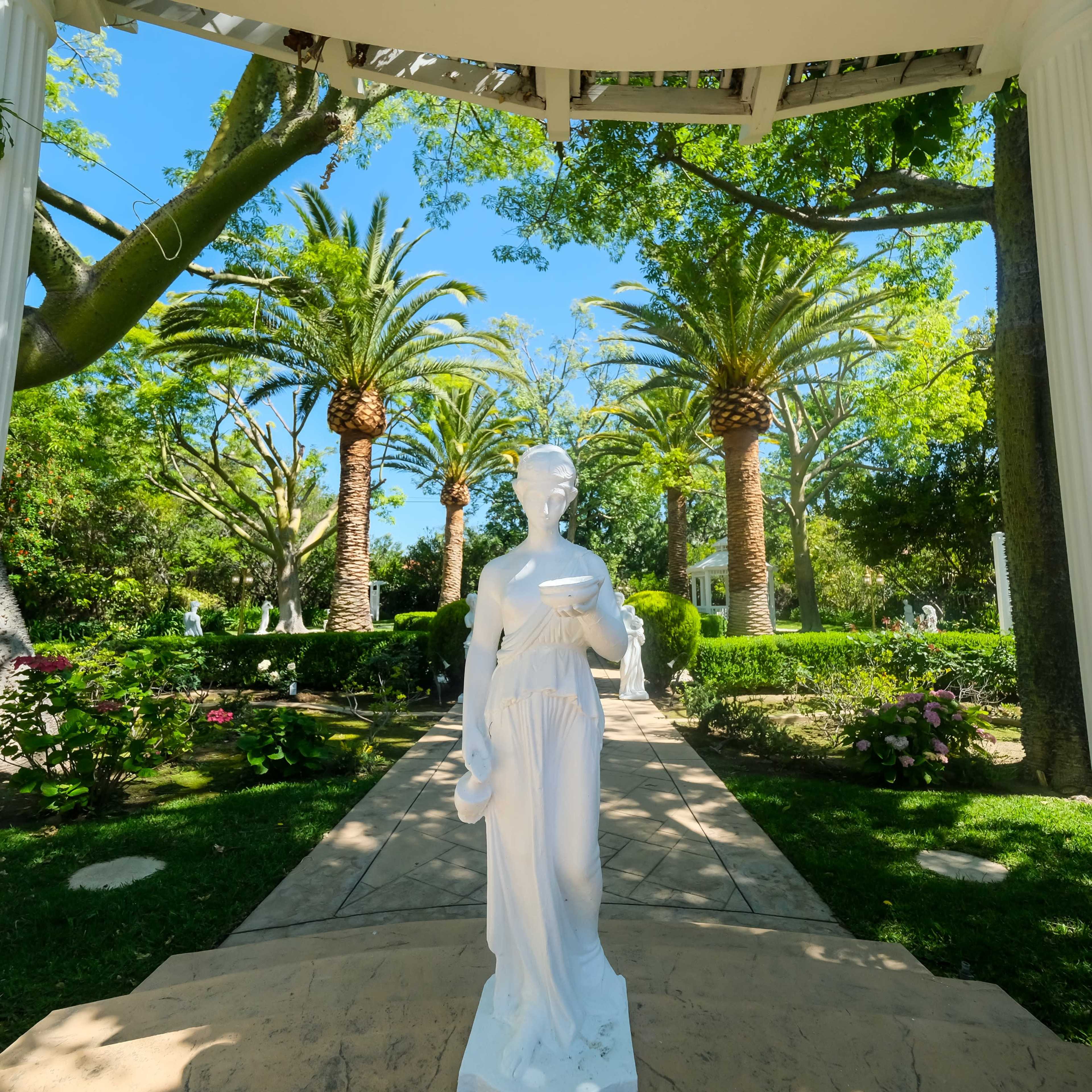 A white statue stands at the center of a garden framed by tall palm trees and lush greenery, with a stone path leading deeper into the landscape.