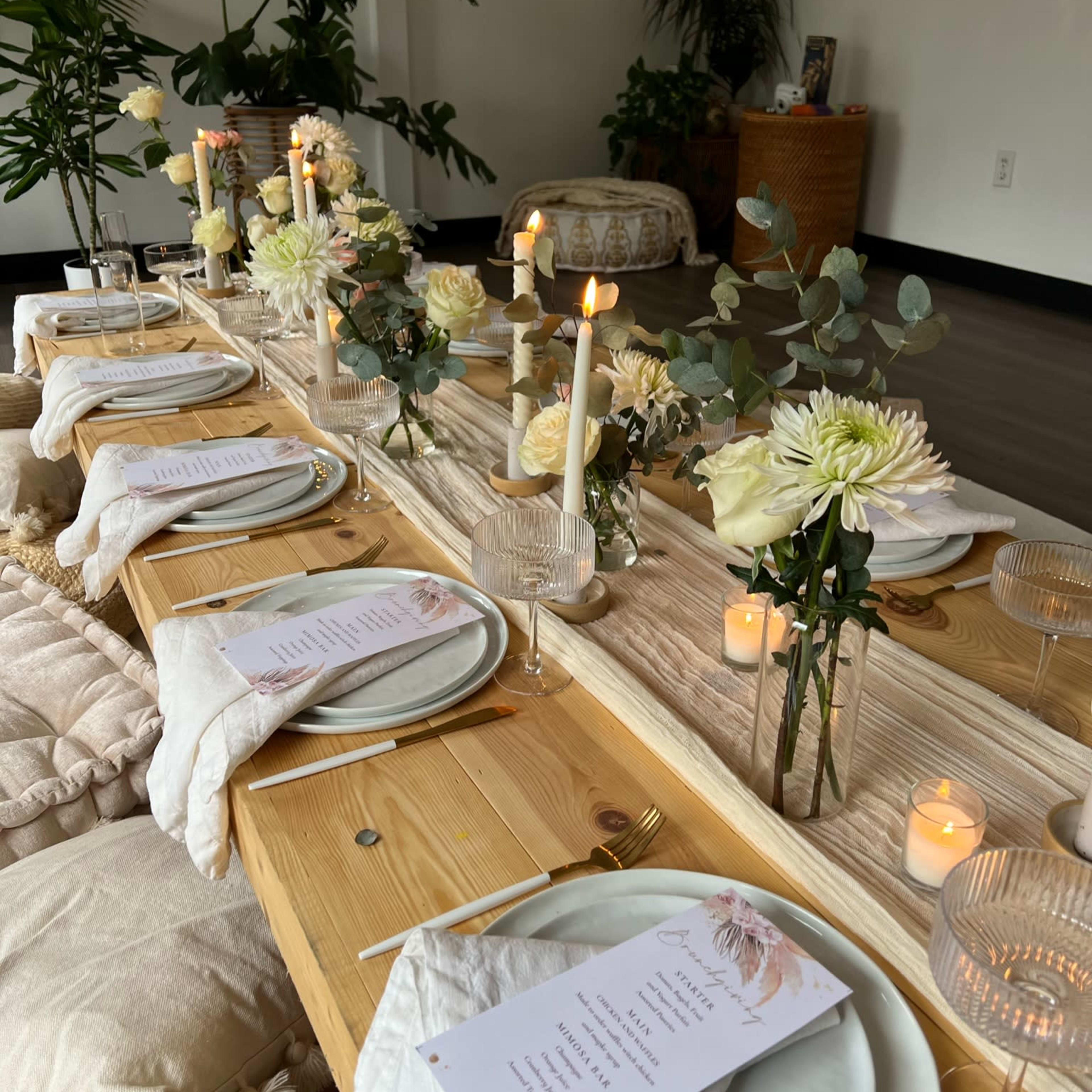 A long wooden table is set for a dinner, adorned with white flowers, candles, and elegant place settings.