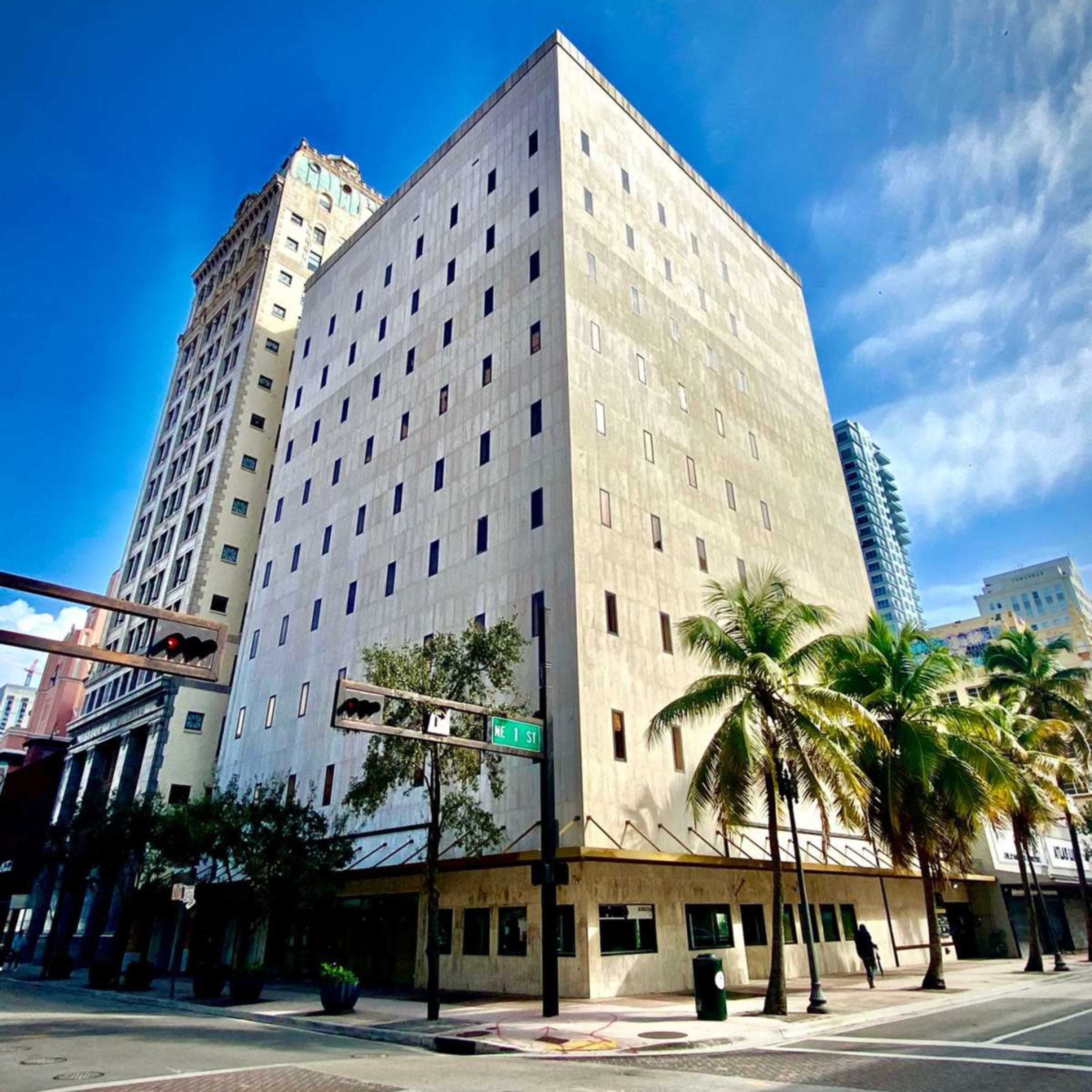 A modern, rectangular concrete building stands next to palm trees along a city street under a clear blue sky.